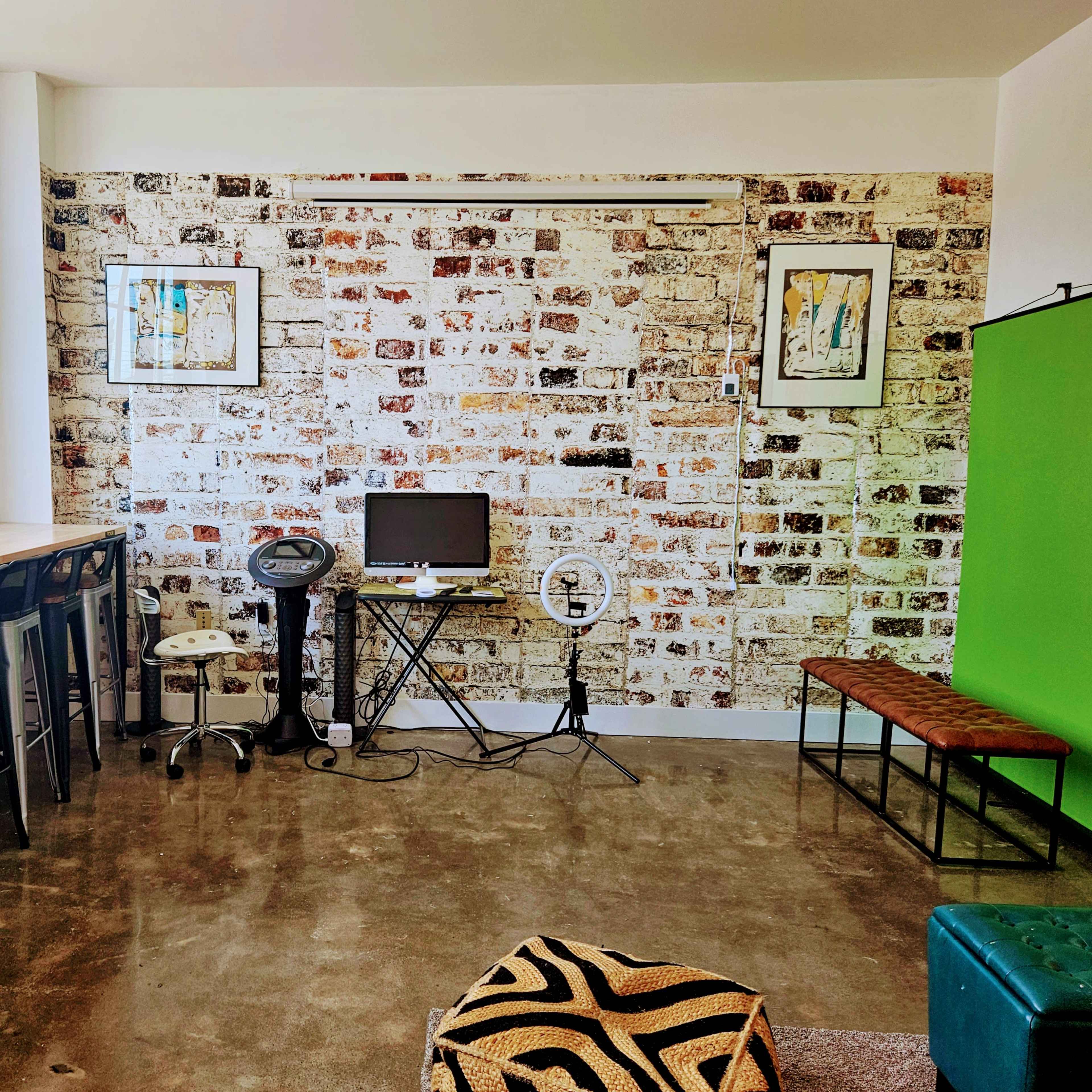 A room with a brick wall, a small desk with a monitor, a ring light on a stand, barstools along a countertop, and a green screen backdrop.