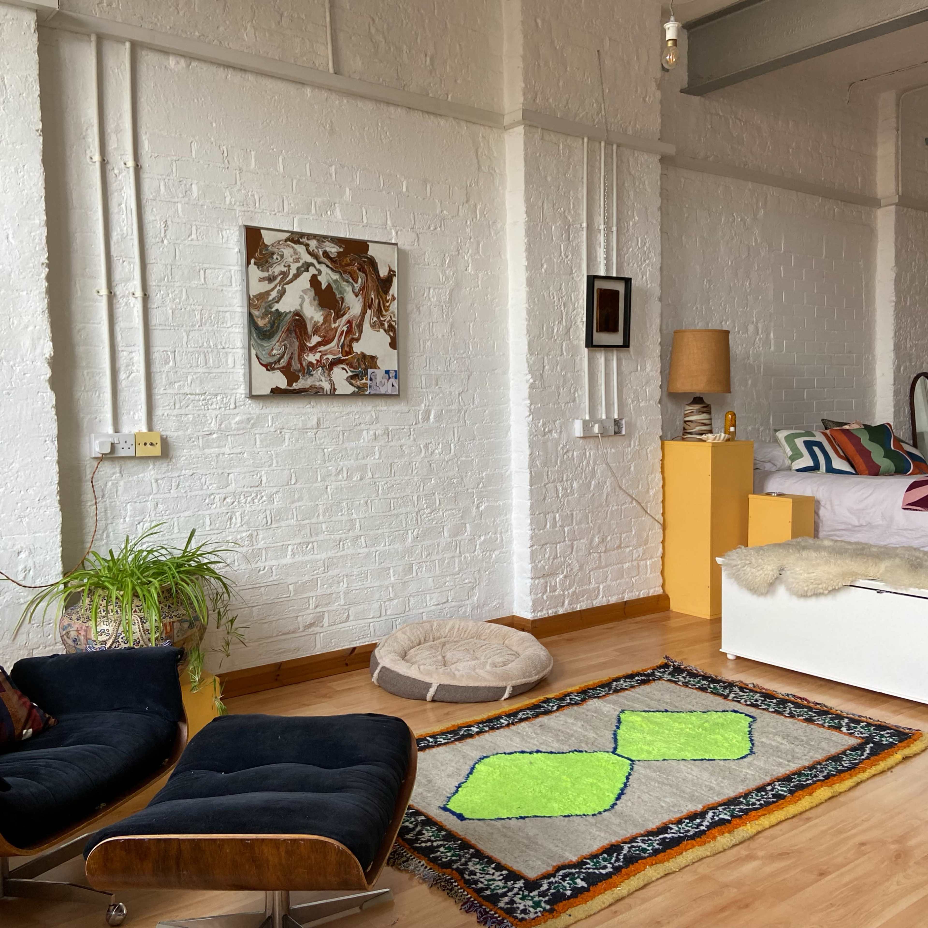 A bright, airy studio apartment features a bed, a black armchair, a colorful rug with green accents, and artwork on the white brick walls.