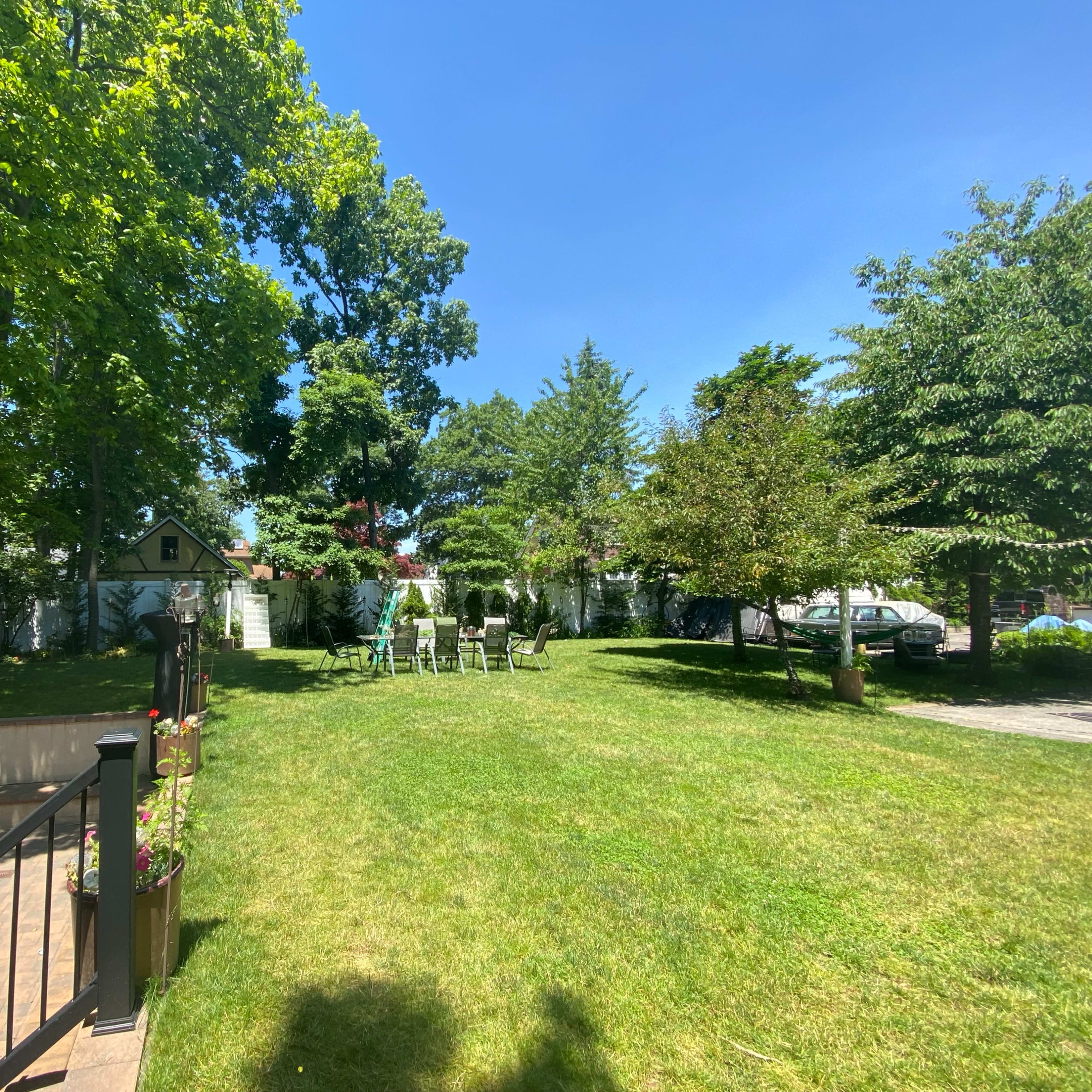 A spacious backyard features green grass, several trees, and a set of chairs arranged under the sunlight.
