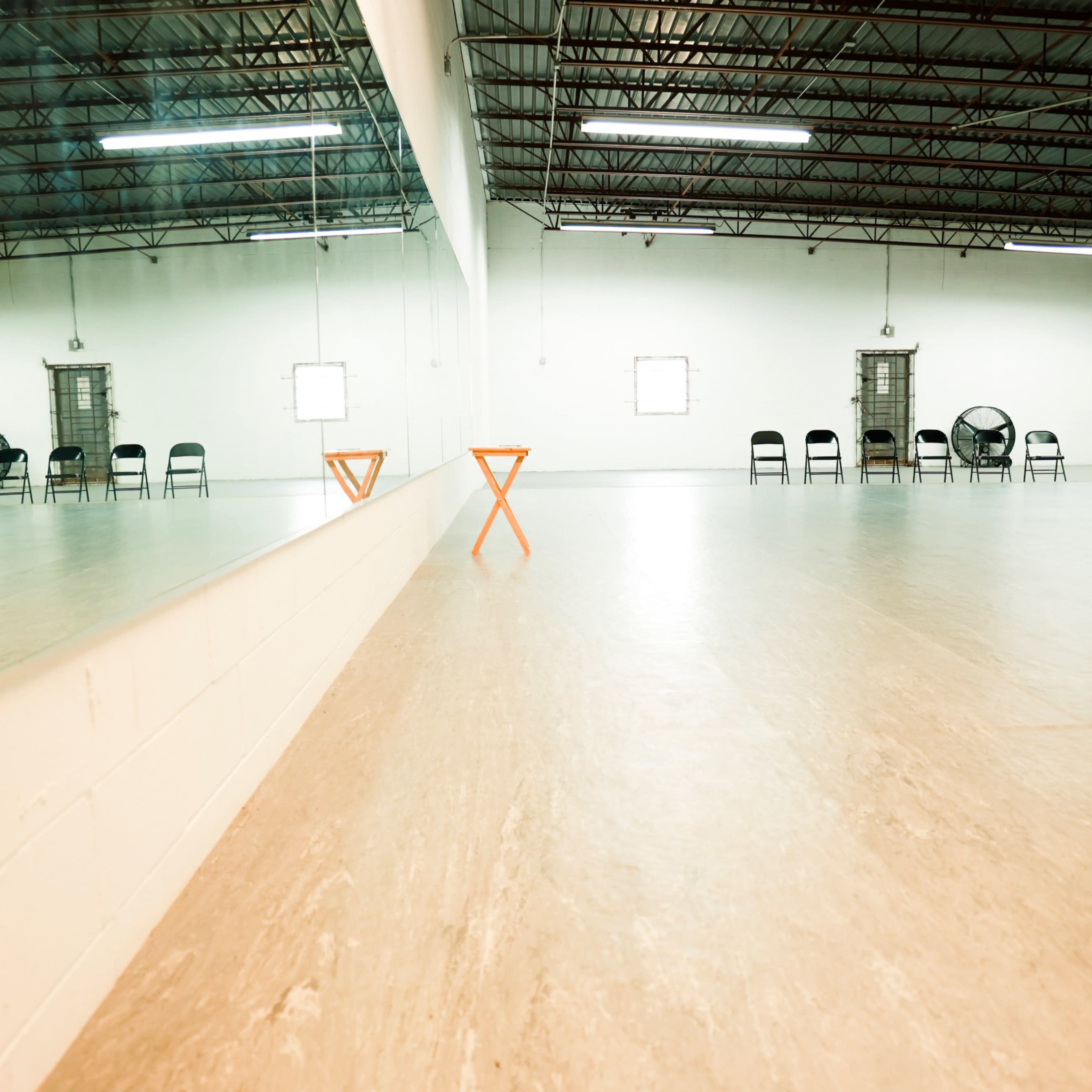 An empty room with wooden flooring, large mirrors on one wall, and a row of black chairs and fans along the opposite side.