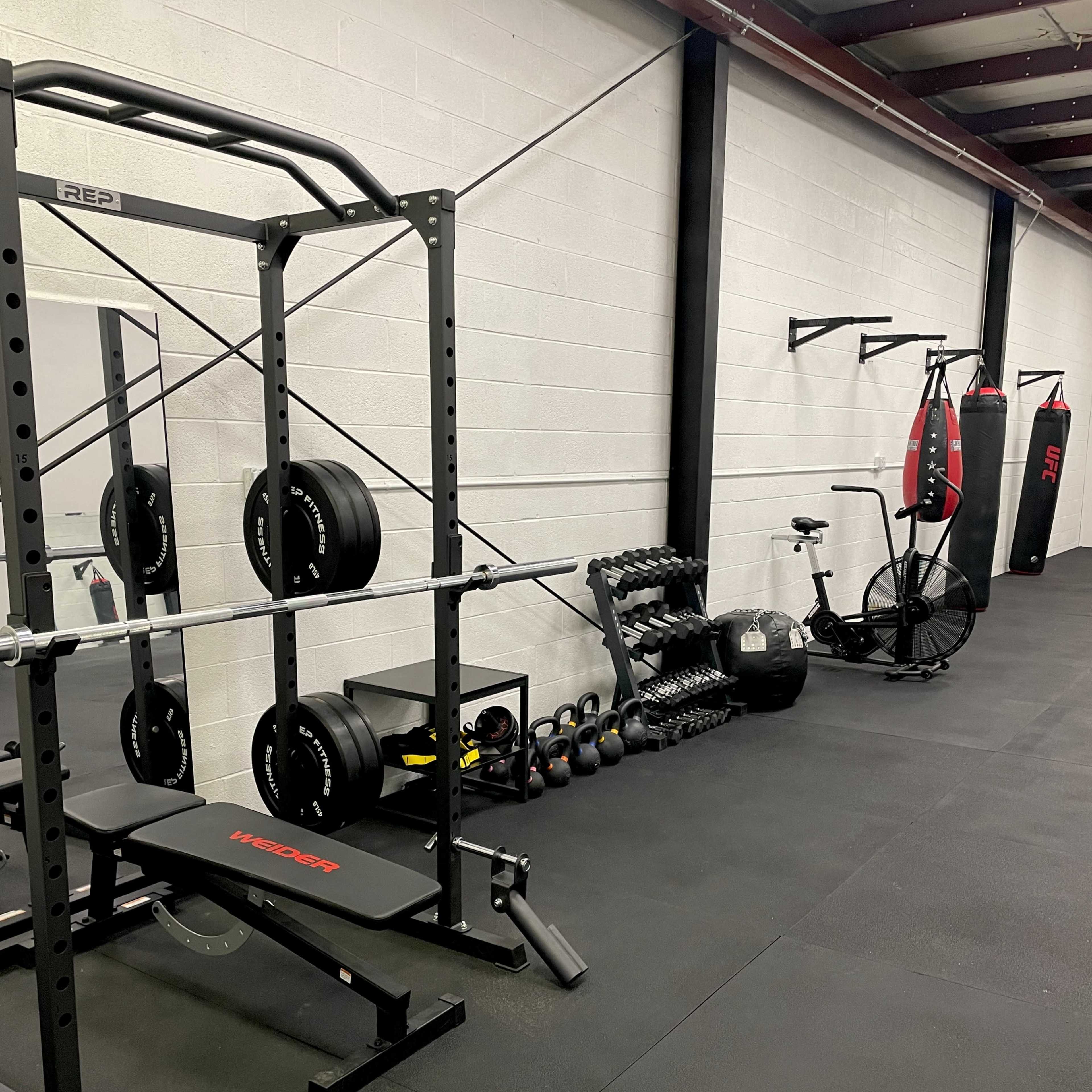 A gym interior featuring a squat rack with weights, a bench, a rowing machine, a wall-mounted rack, and a stationary bike.