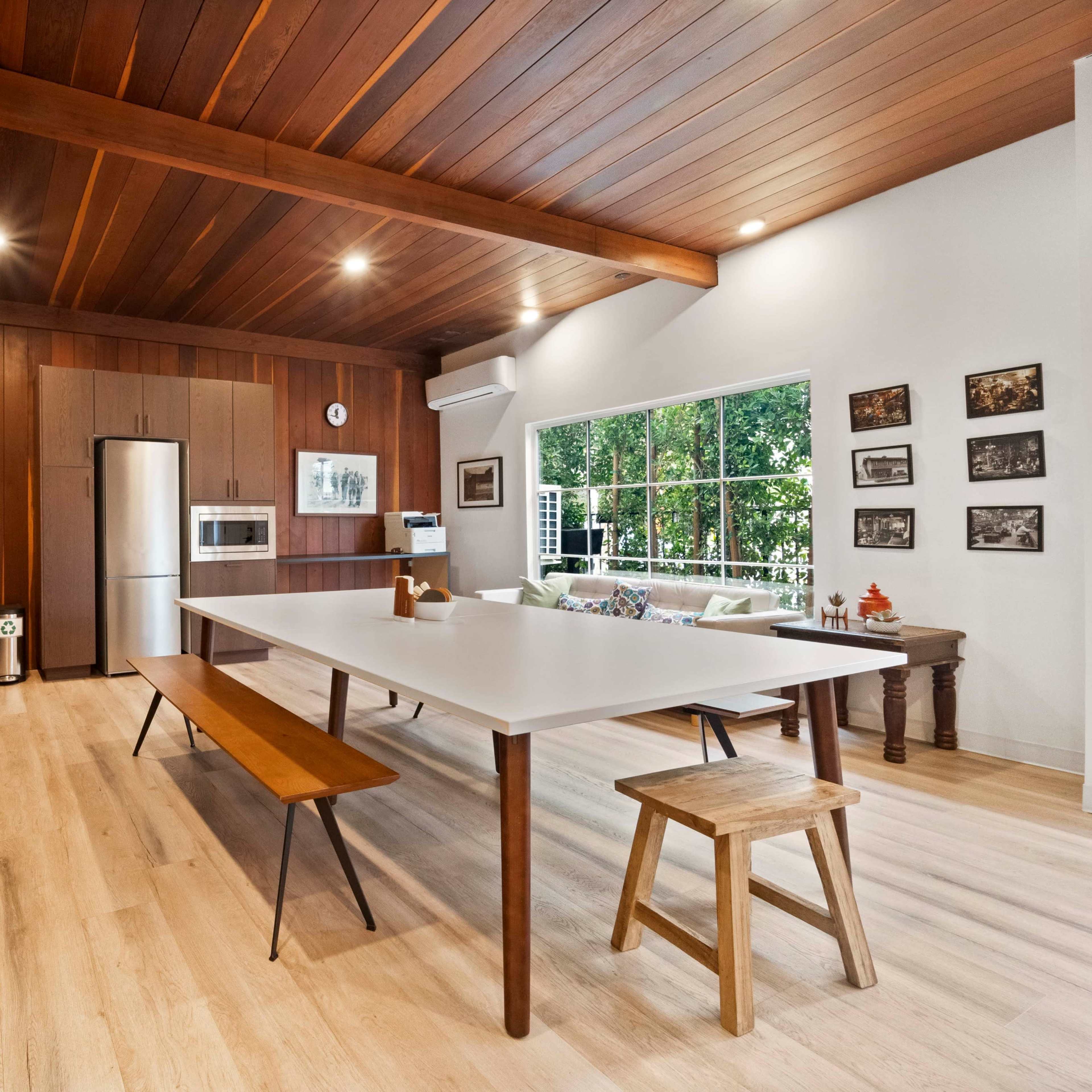 A modern kitchen features a large white table, wooden cabinetry, and a view of greenery through a large window.