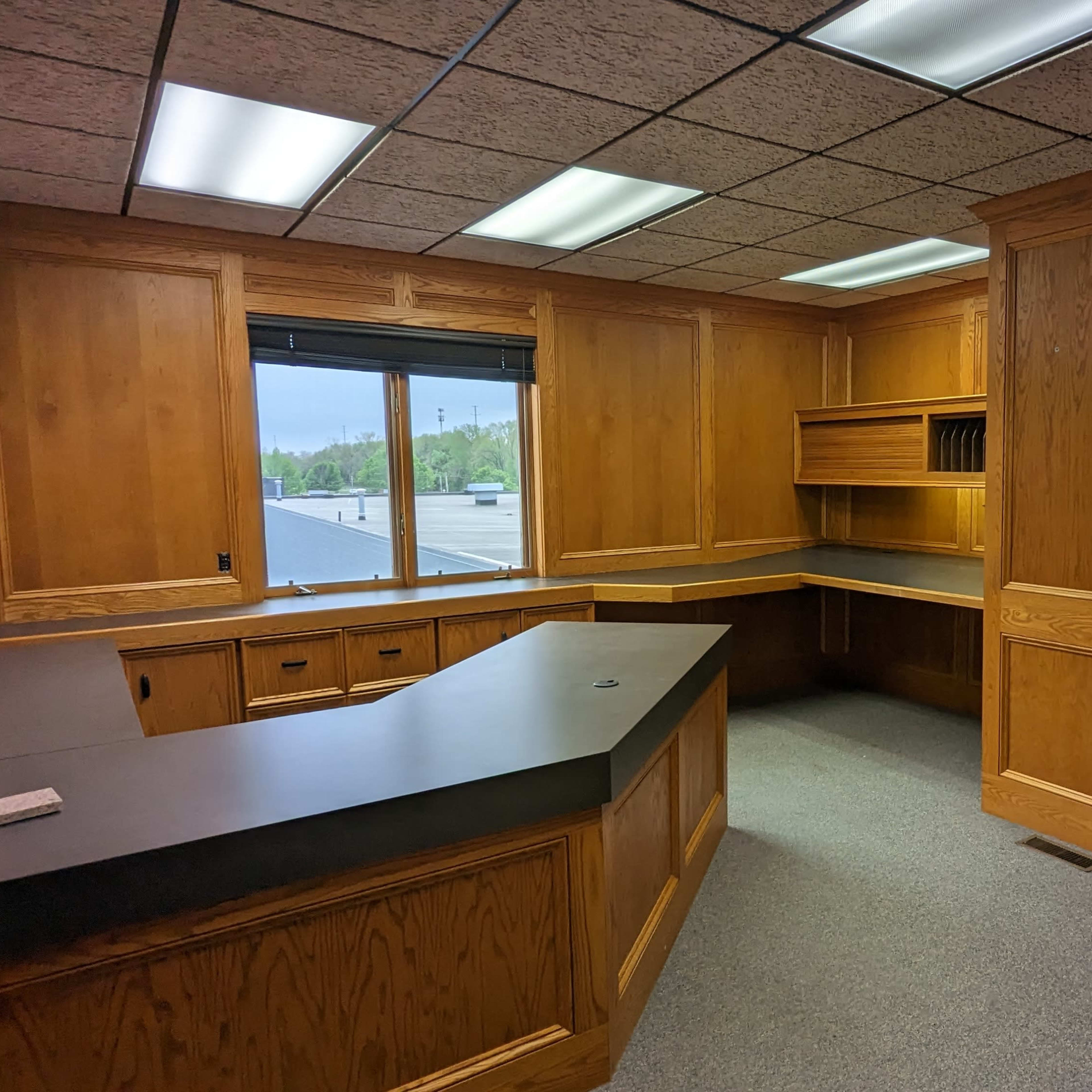 A vacant office space with wooden paneling, a large desk, and a window overlooking an external view.