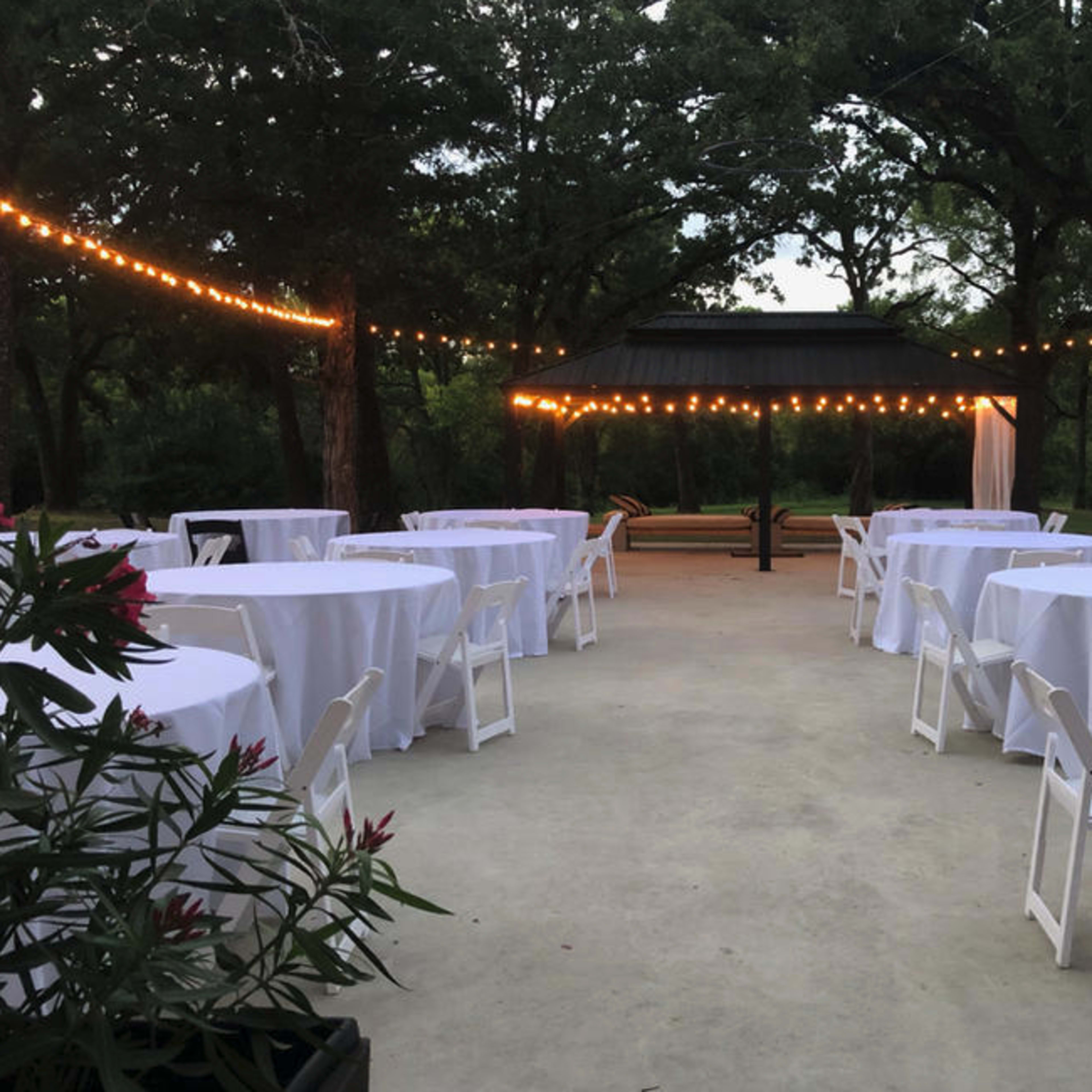 A landscaped outdoor area is set up for an event with round tables covered in white linens and string lights overhead.