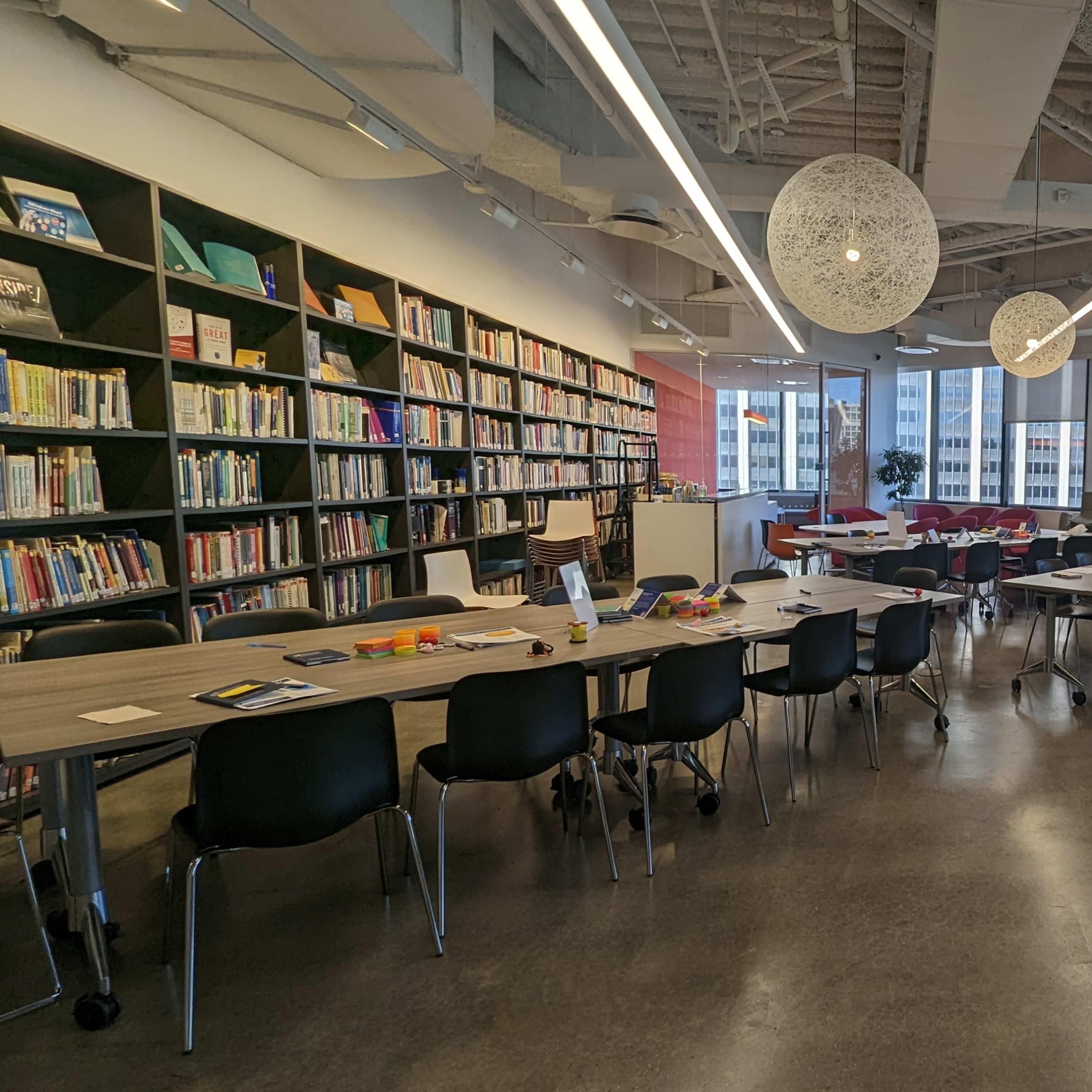 A modern meeting space with long tables arranged in a U-shape, surrounded by shelves filled with books and large windows letting in natural light.