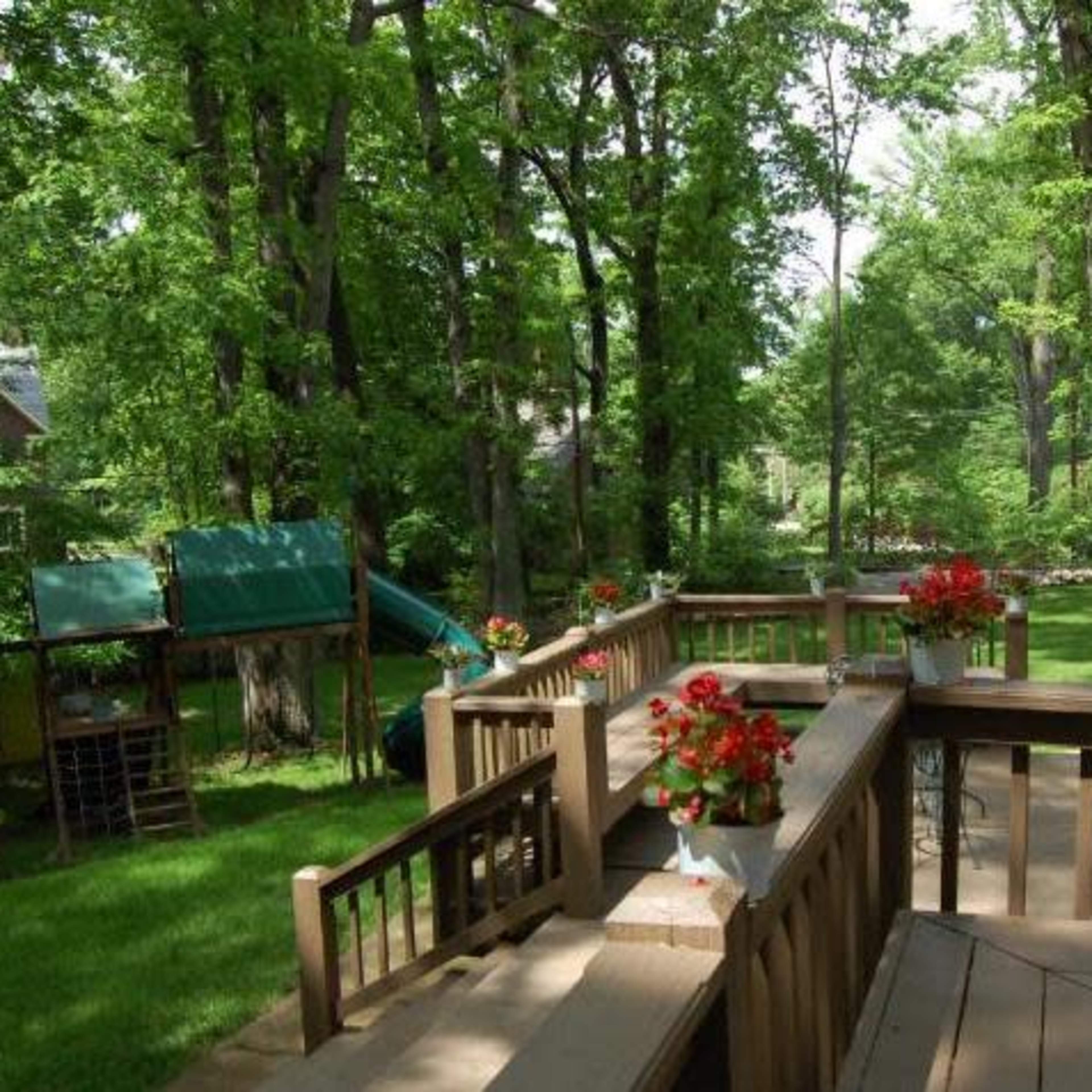 A wooden deck surrounded by lush green trees features potted flowers and outdoor seating areas.
