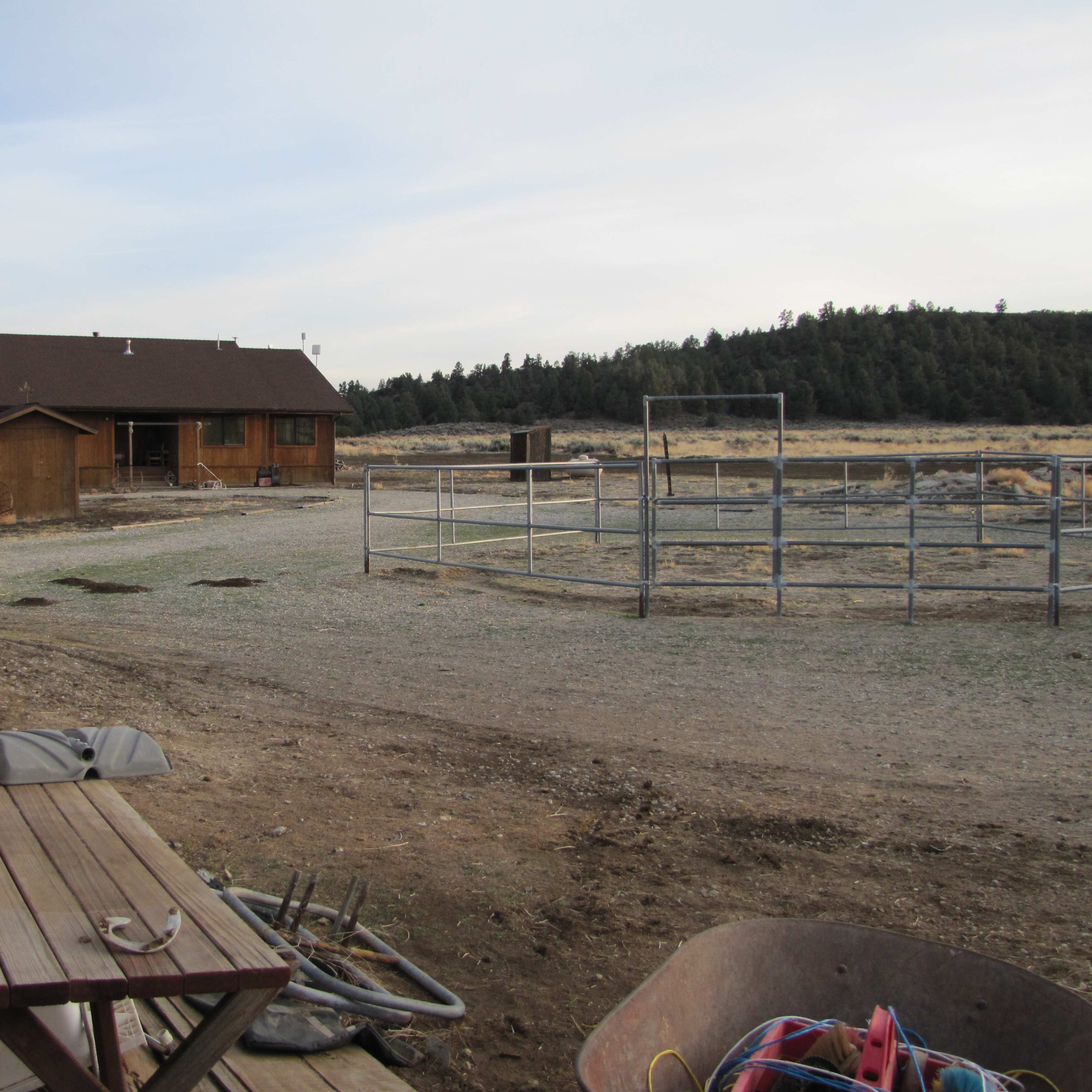 A wooden house is situated near a fenced corral in a rural landscape with sparse vegetation and mountains in the distance.