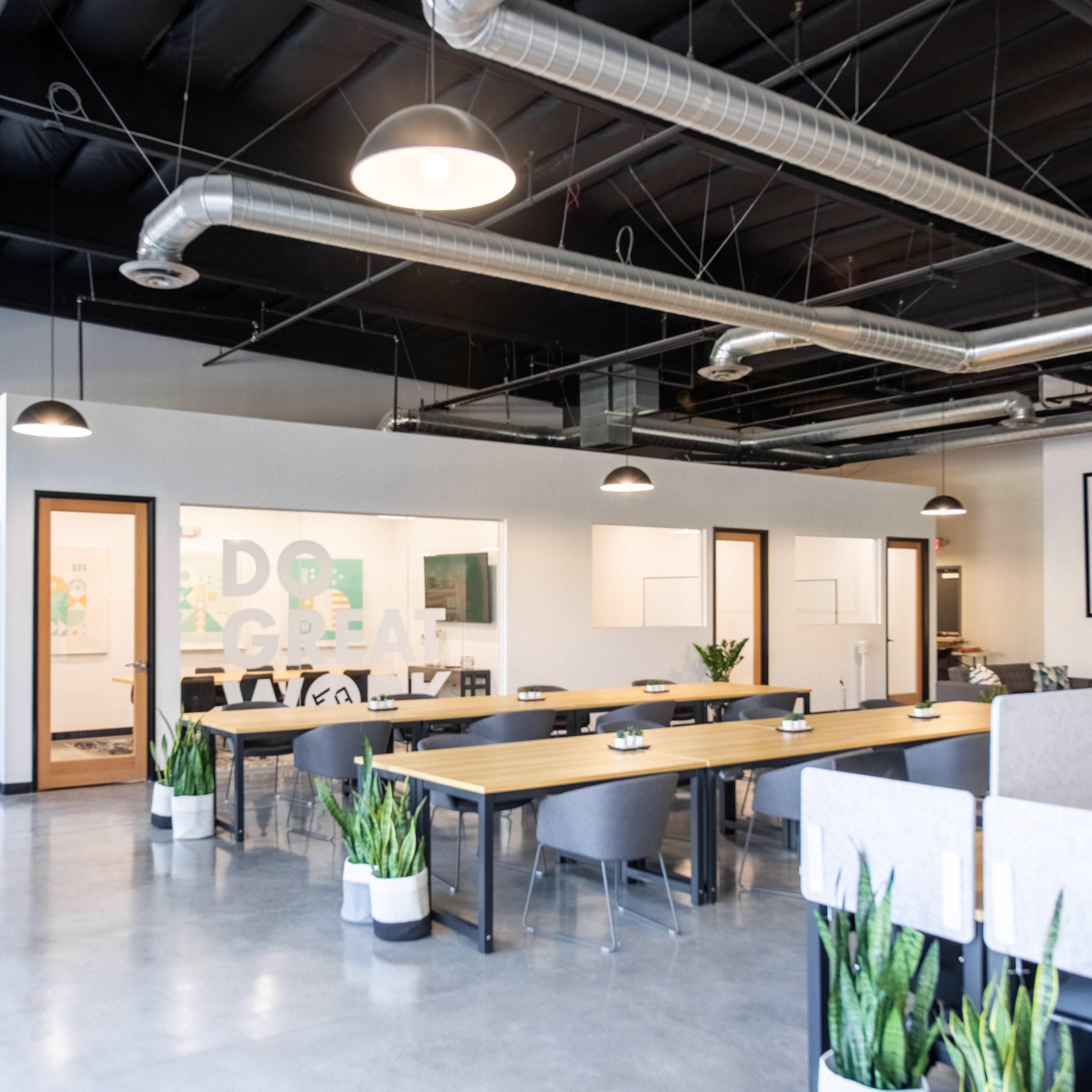 A modern office space featuring a large communal table, workstation dividers, and meeting rooms with glass walls, surrounded by potted plants.