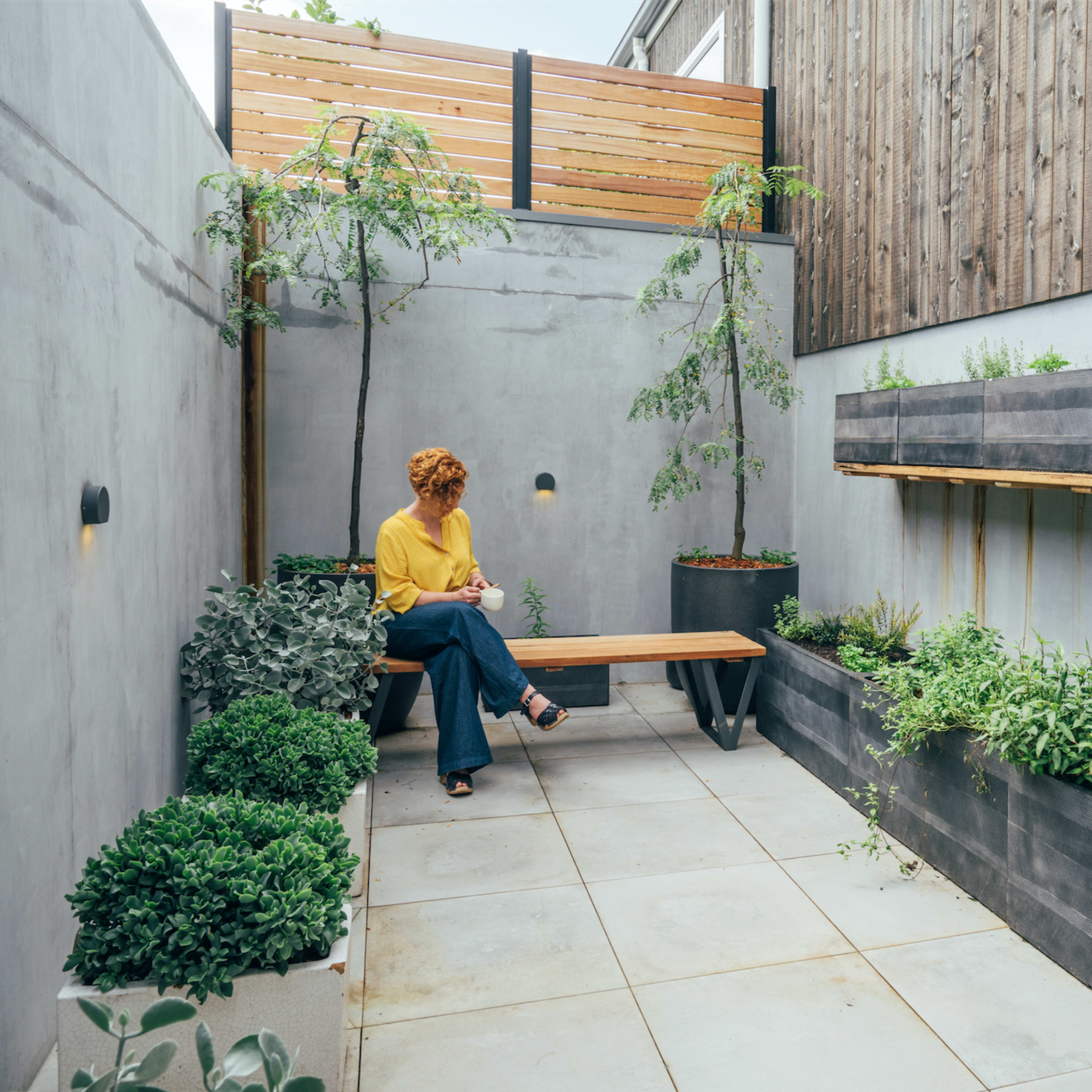 A person sits on a wooden bench in a narrow, modern garden space surrounded by concrete walls and raised planters filled with greenery.