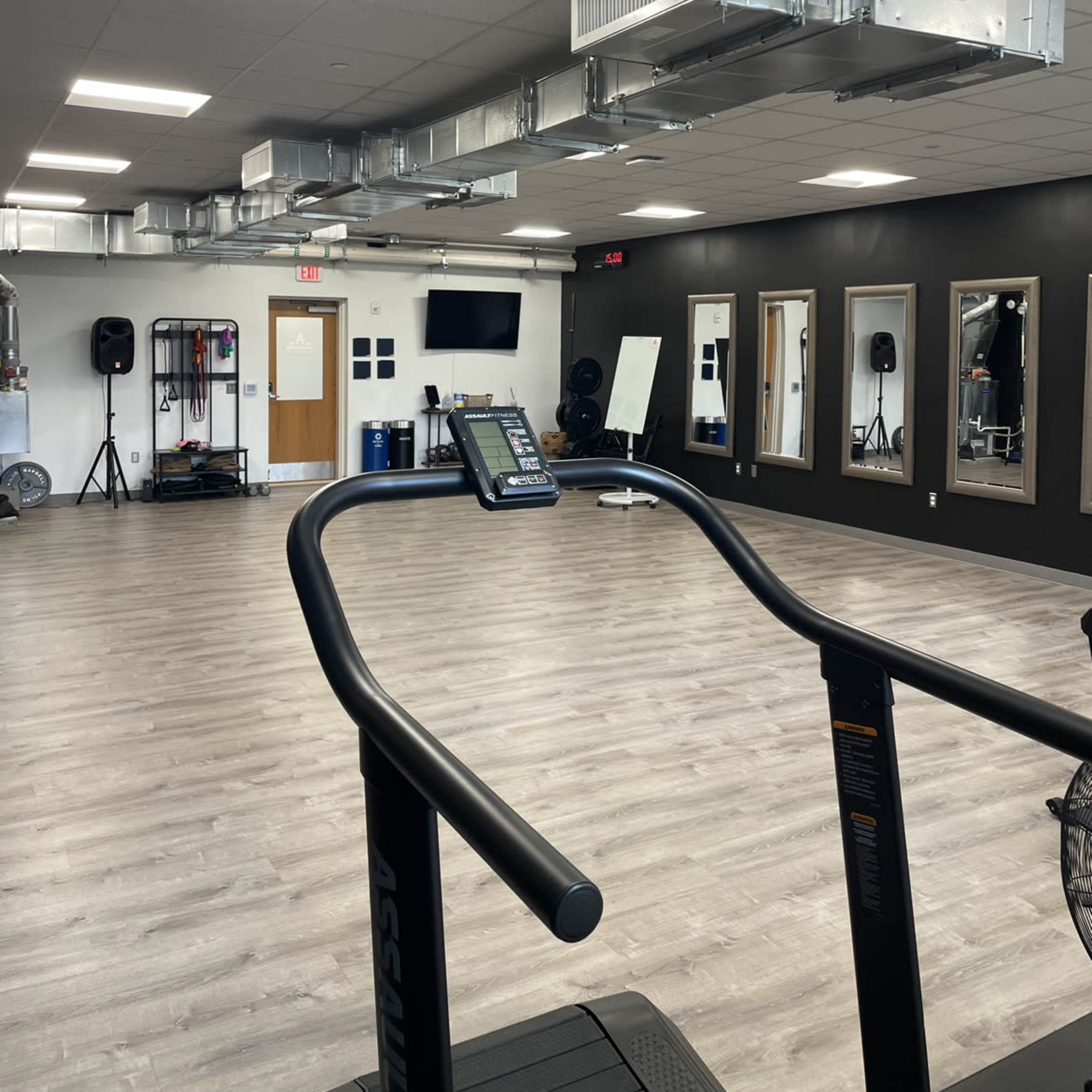 A modern gym interior with a treadmill in the foreground, large mirrors on the walls, and ventilation ducts on the ceiling.
