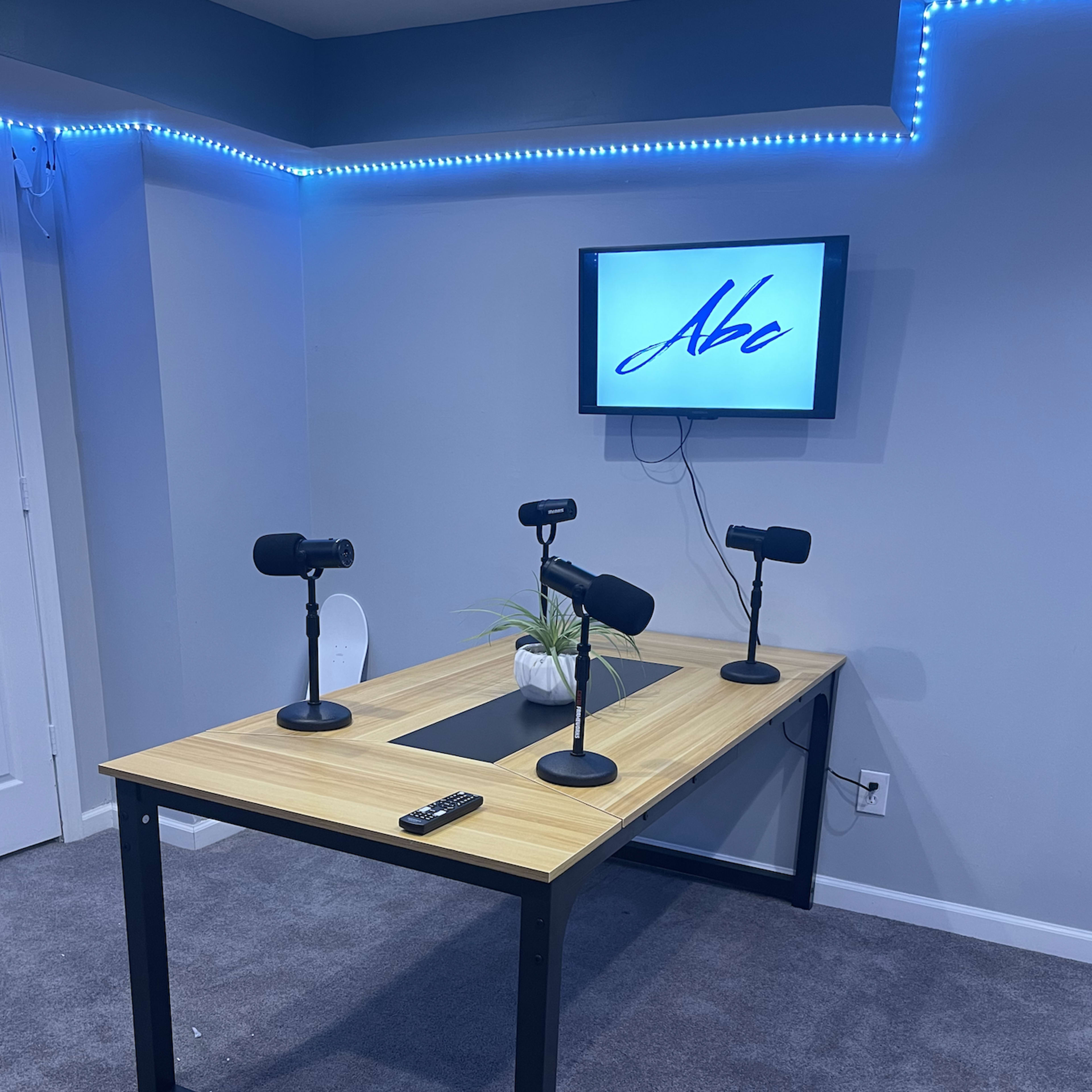 A podcasting setup with a wooden table, three microphones on stands, a small plant, and a screen displaying the text "Aba" mounted on a wall.