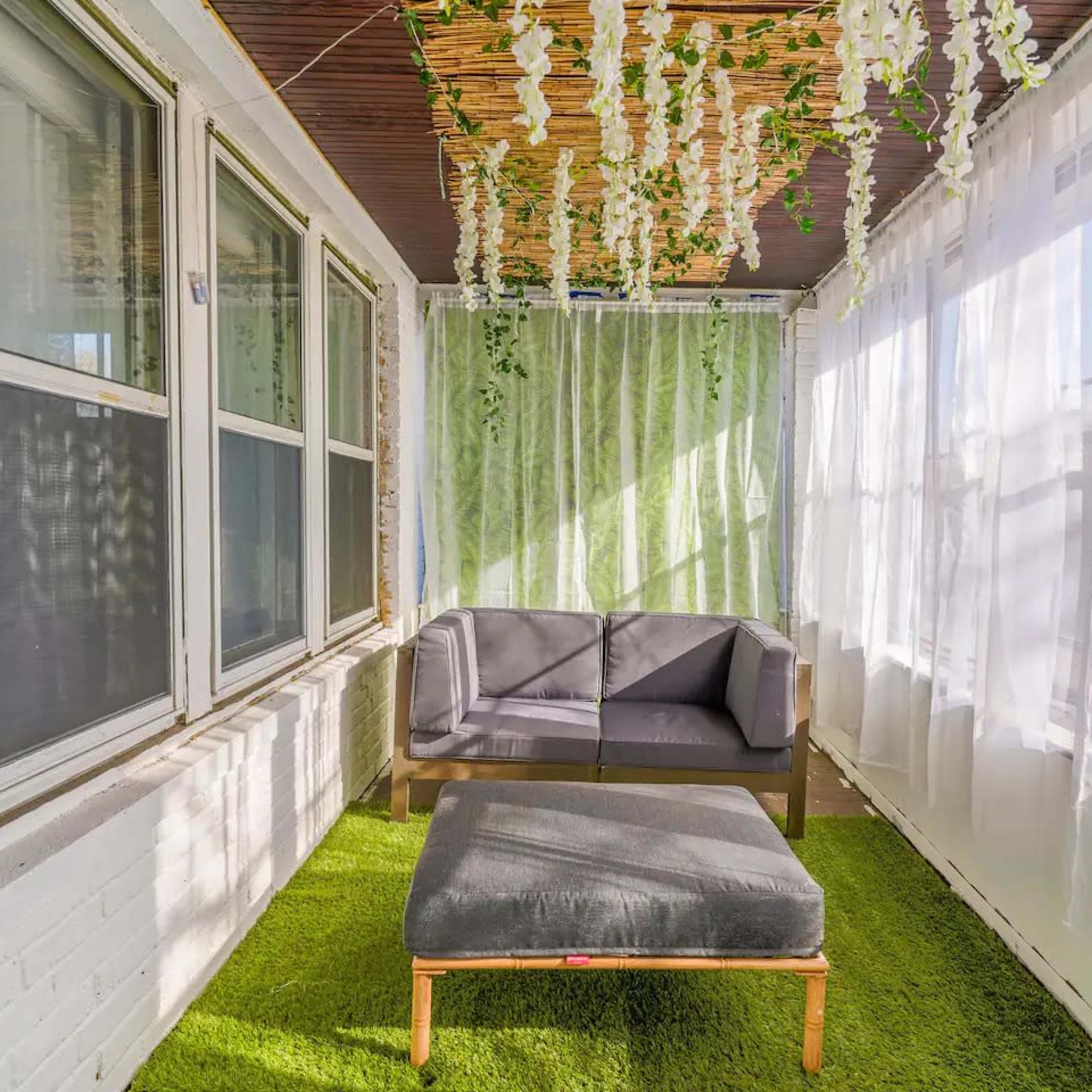 A sunlit sunroom features a gray sofa and ottoman on green carpet, surrounded by sheer curtains and hanging greenery.