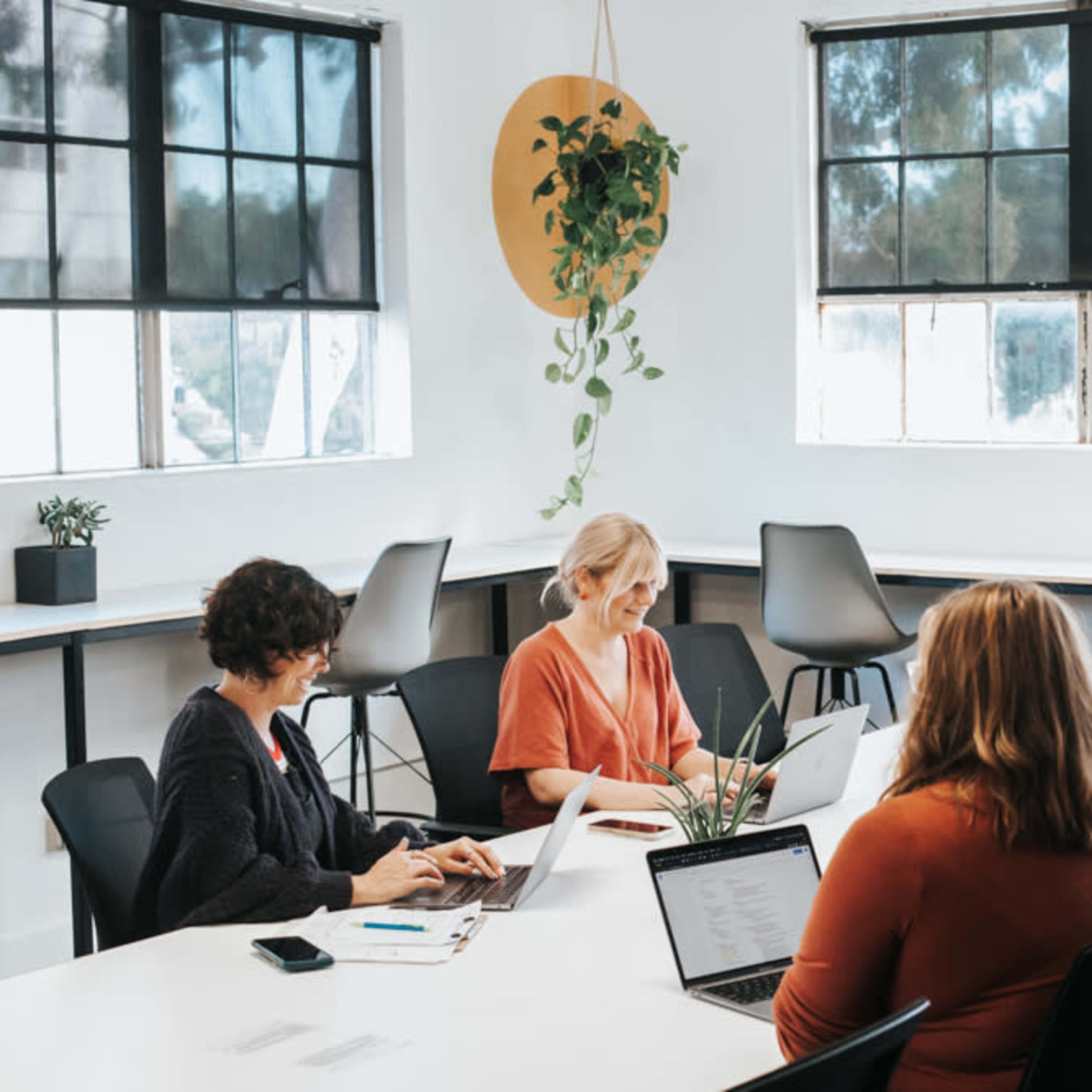 A group of five people are seated around a table in a well-lit office space, working on laptops and notebooks.