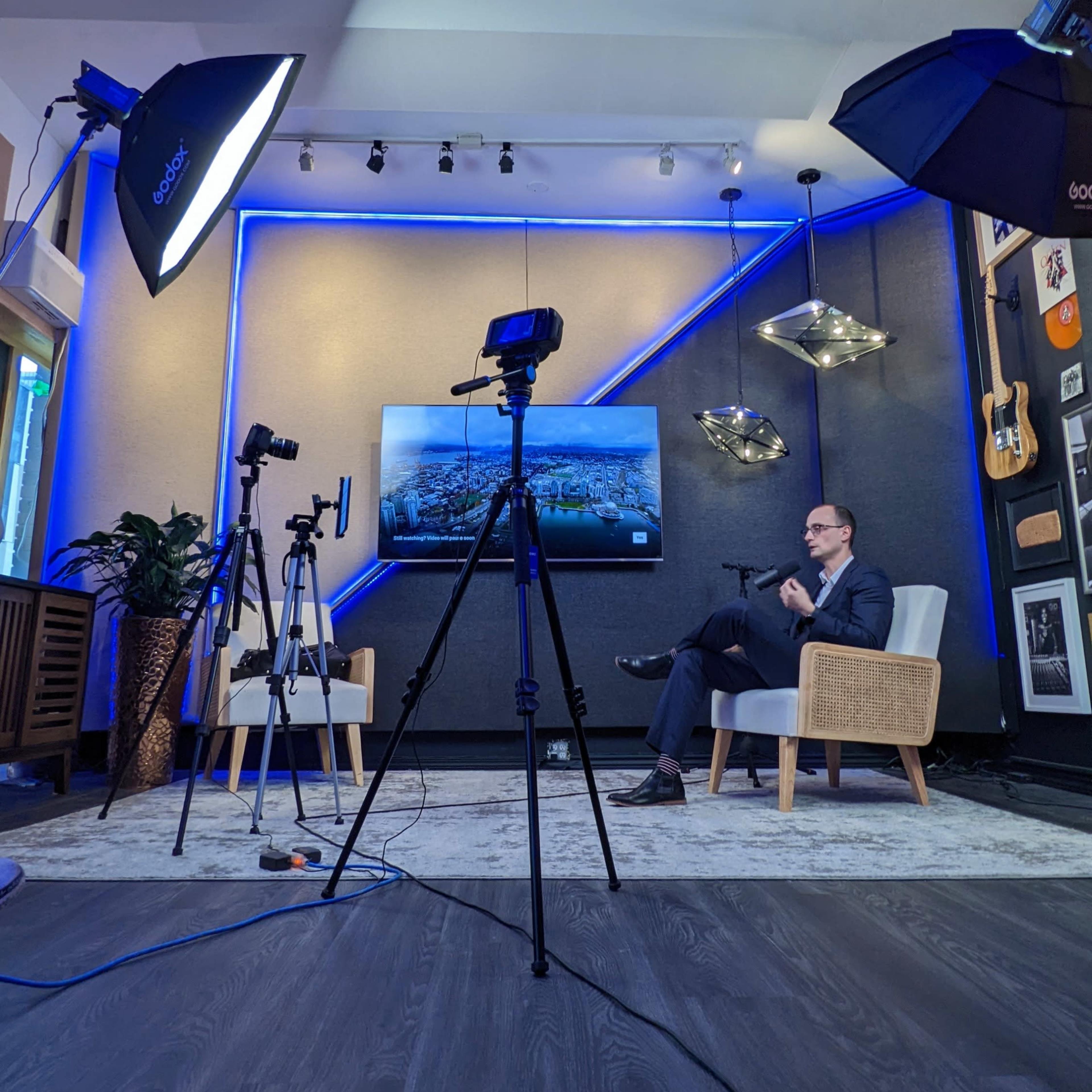 A professionally arranged studio setup with cameras and lighting focused on a seated speaker in front of a large screen displaying a cityscape.