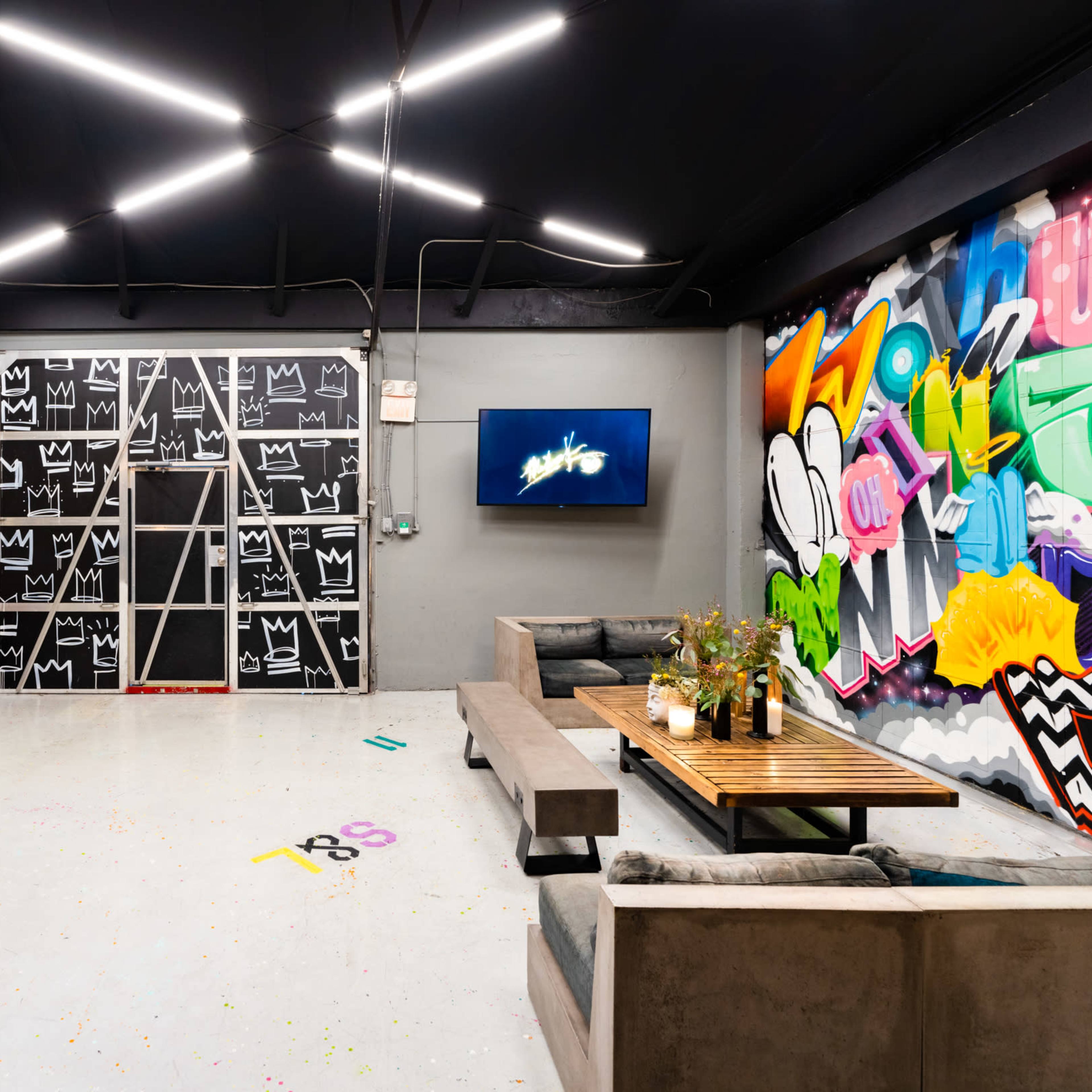 A modern, open space featuring a dining table, a lounge area, a colorful mural on one wall, and a large television mounted on another wall.