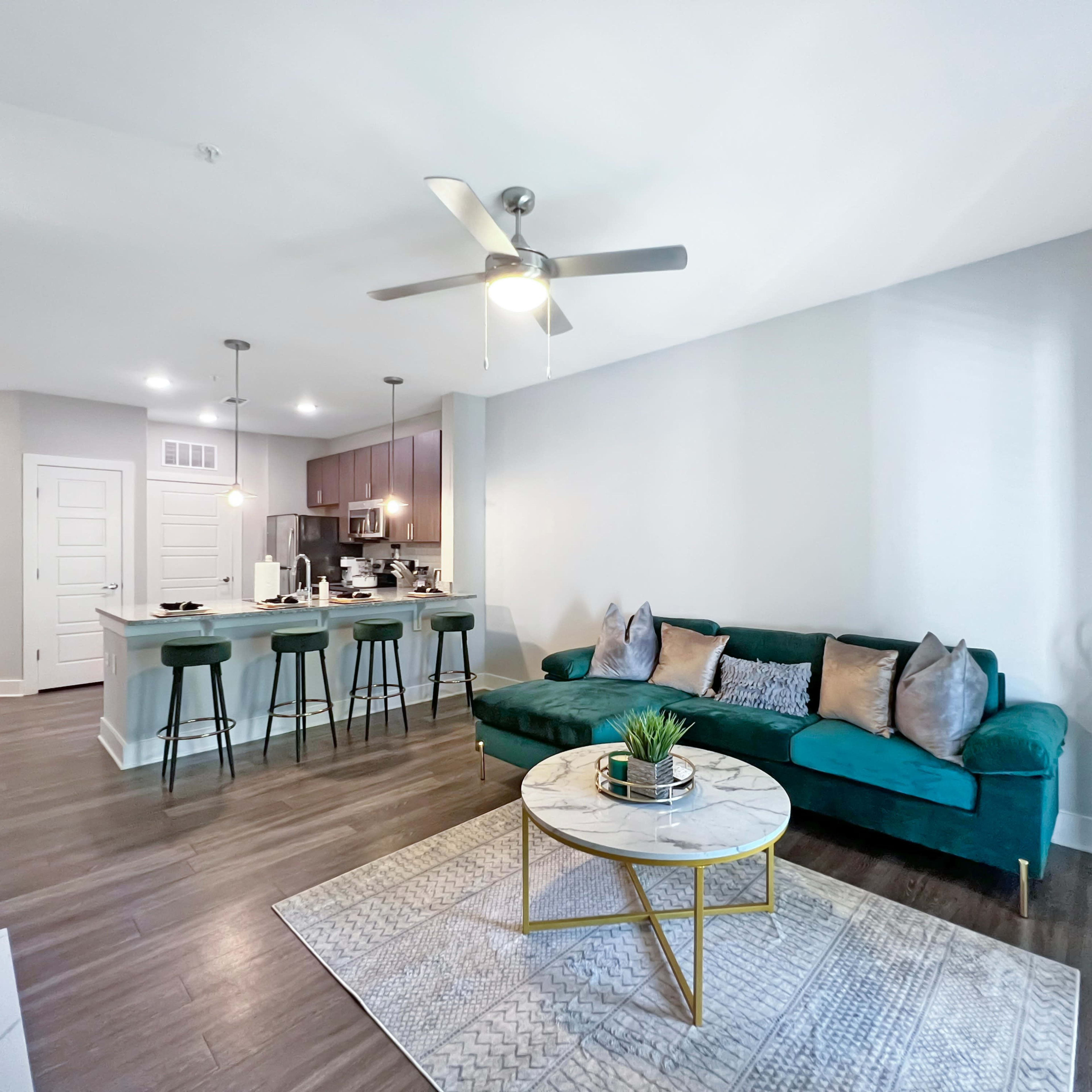 A modern living room with a green sofa, a round coffee table, and a kitchen area featuring bar stools and cabinetry in the background.