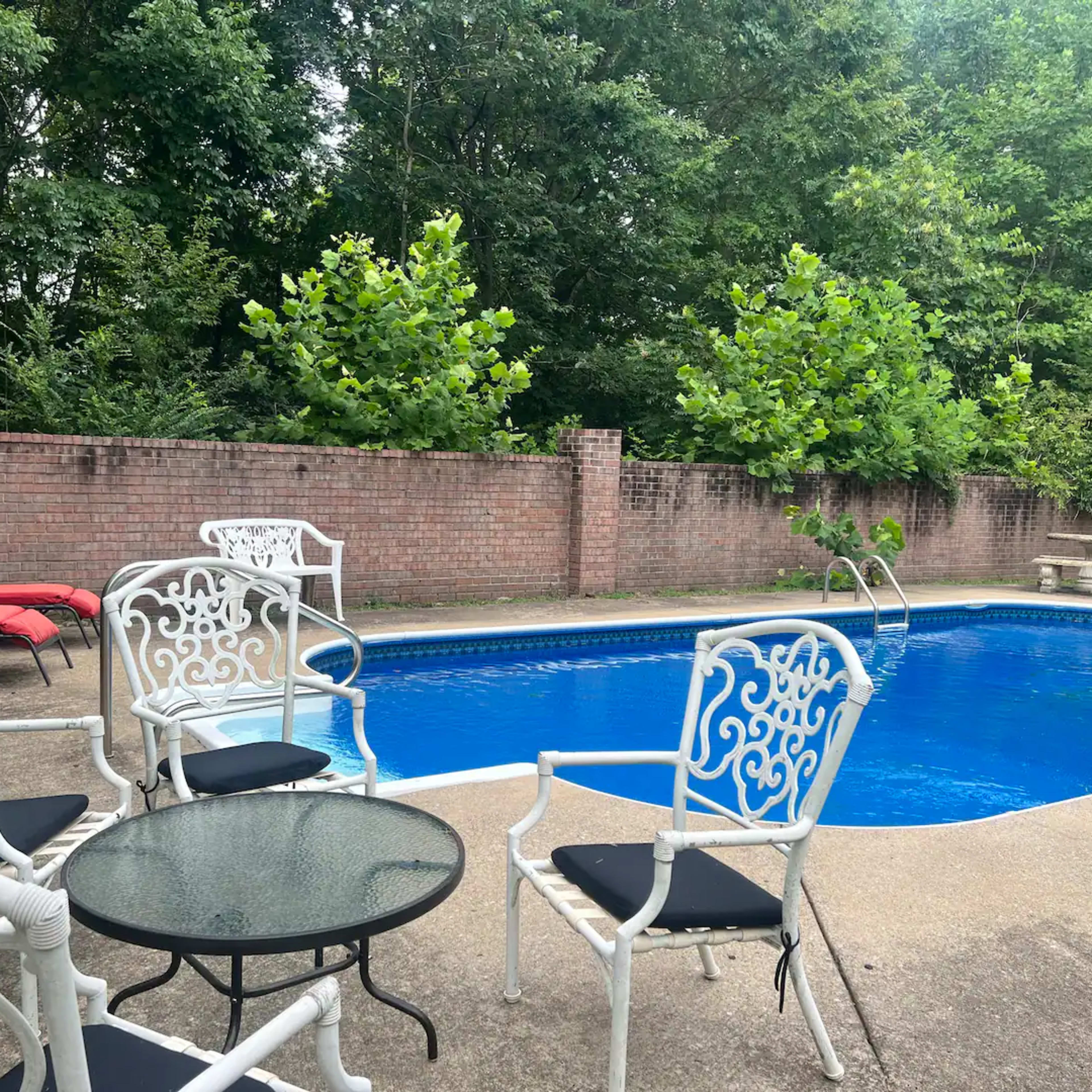 A swimming pool surrounded by white metal chairs and a small table, with a few lounge chairs nearby and greenery in the background.
