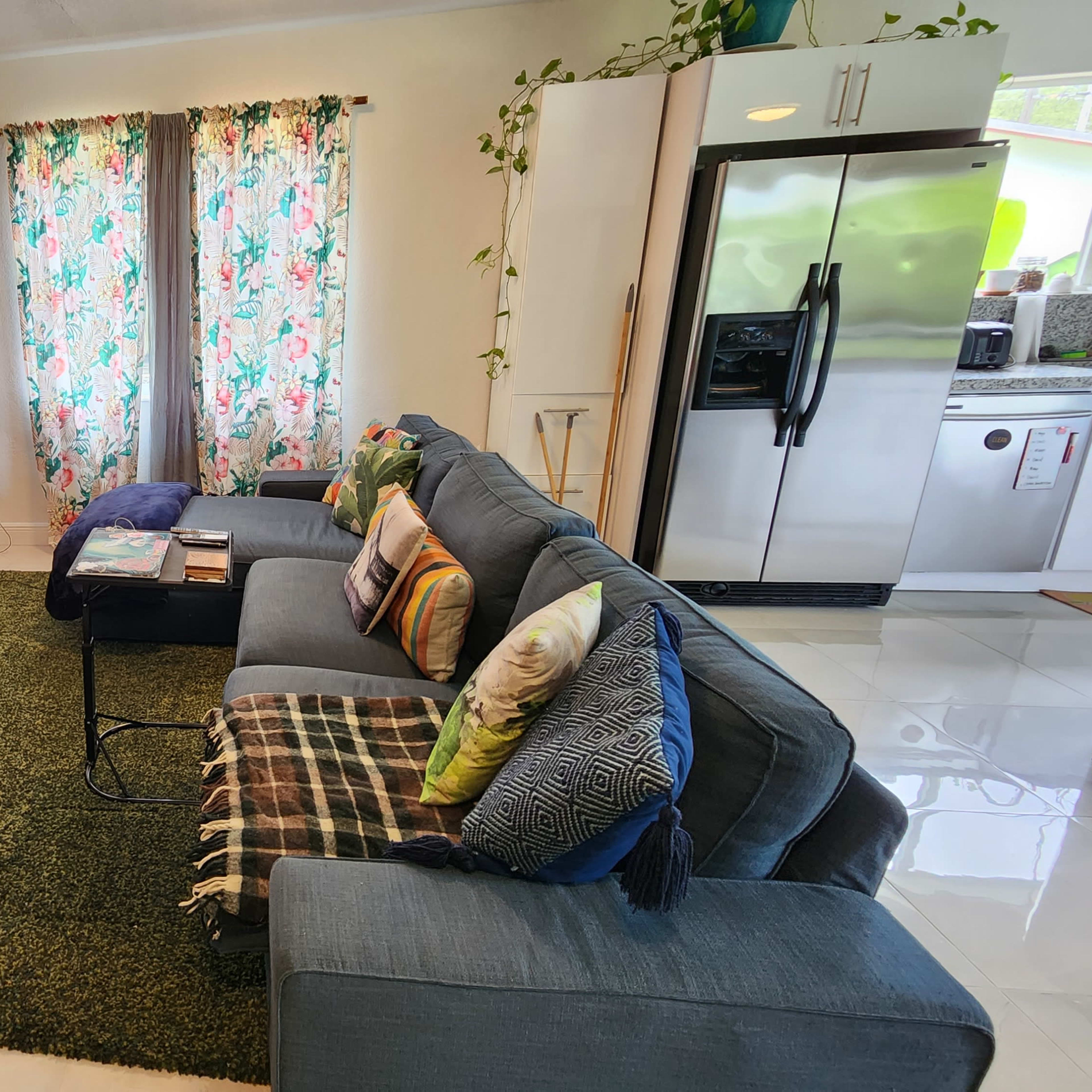 A modern living room features a large sofa with colorful cushions, a TV on a stand, a coffee table, and patterned curtains beside a kitchen area with stainless steel appliances.