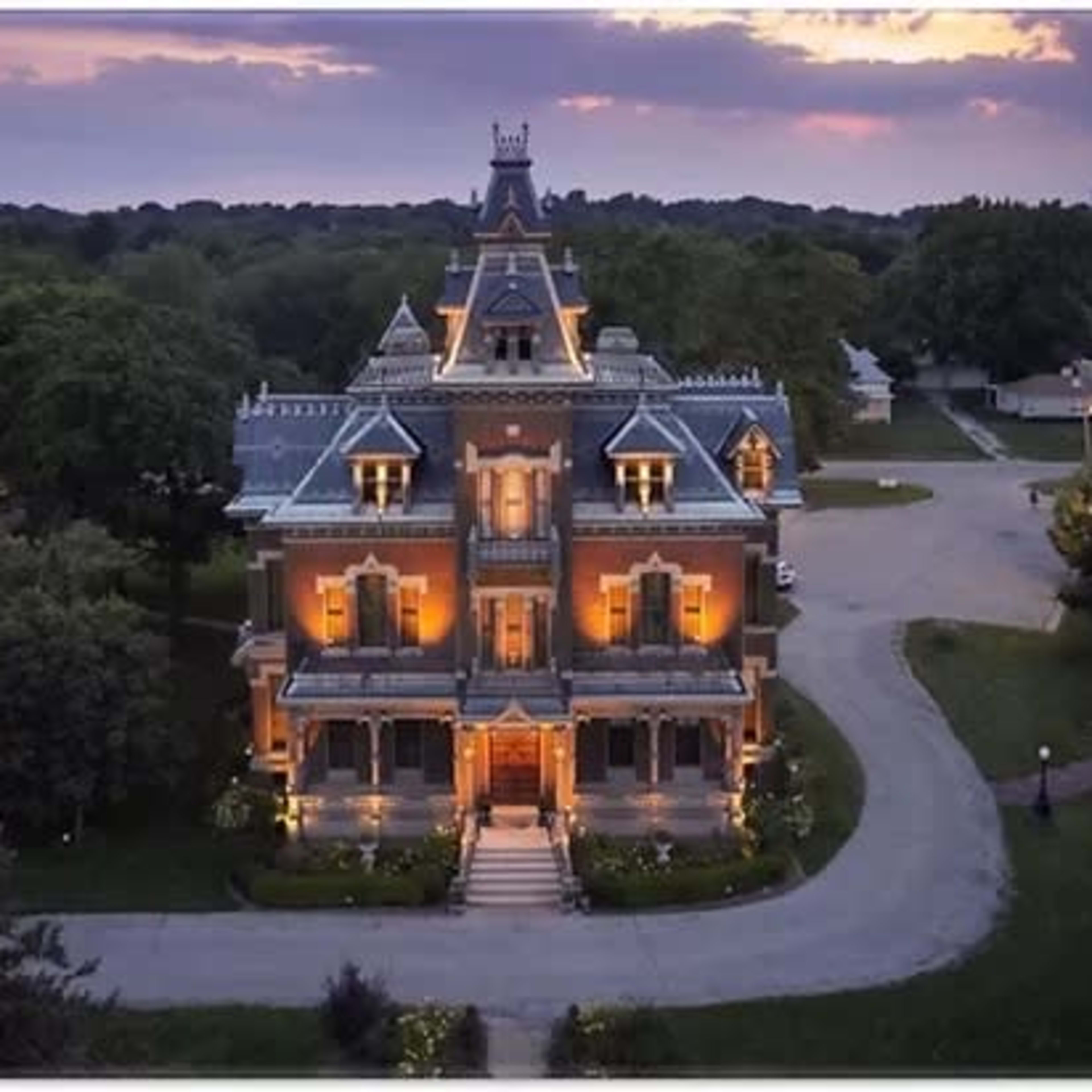 A large, multi-story Victorian mansion with ornate architectural details, illuminated by exterior lighting, set in a tree-lined area at dusk.