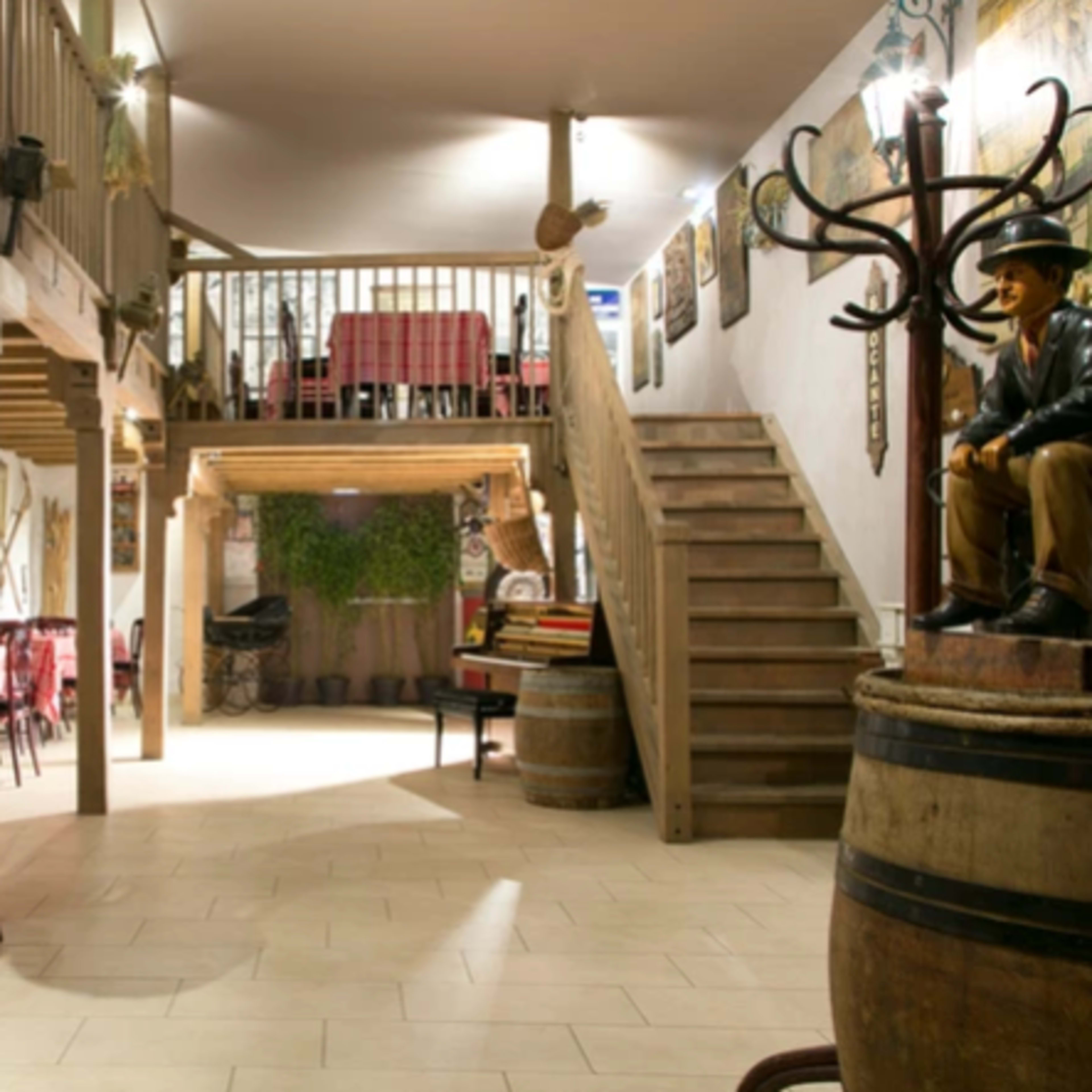 The interior of a rustic restaurant featuring wooden beams, tables with checkered tablecloths, and a staircase leading to an upper level.