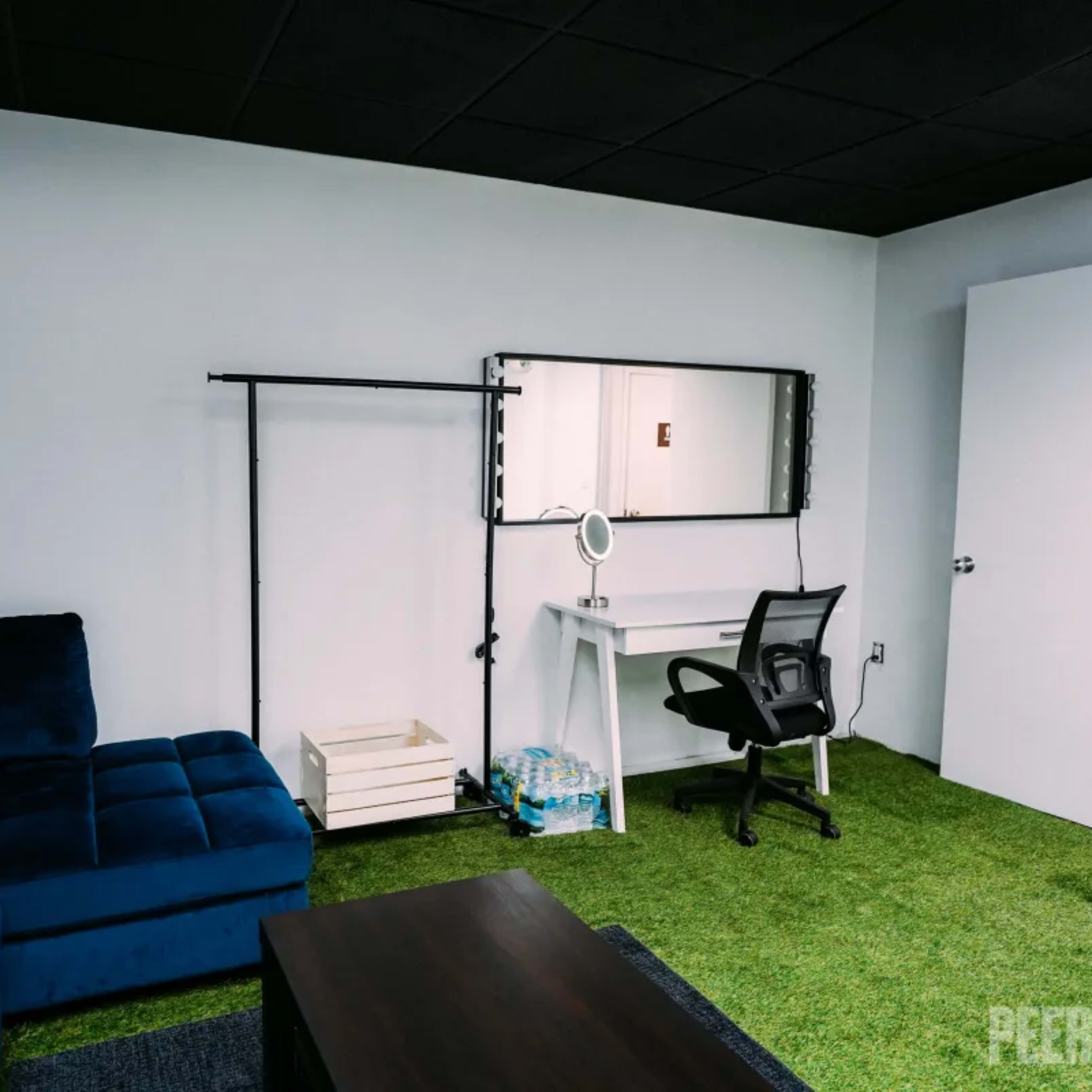 A room with a blue sectional sofa, a white desk with a mirror, a chair, and a rack against the wall, all on green carpet.
