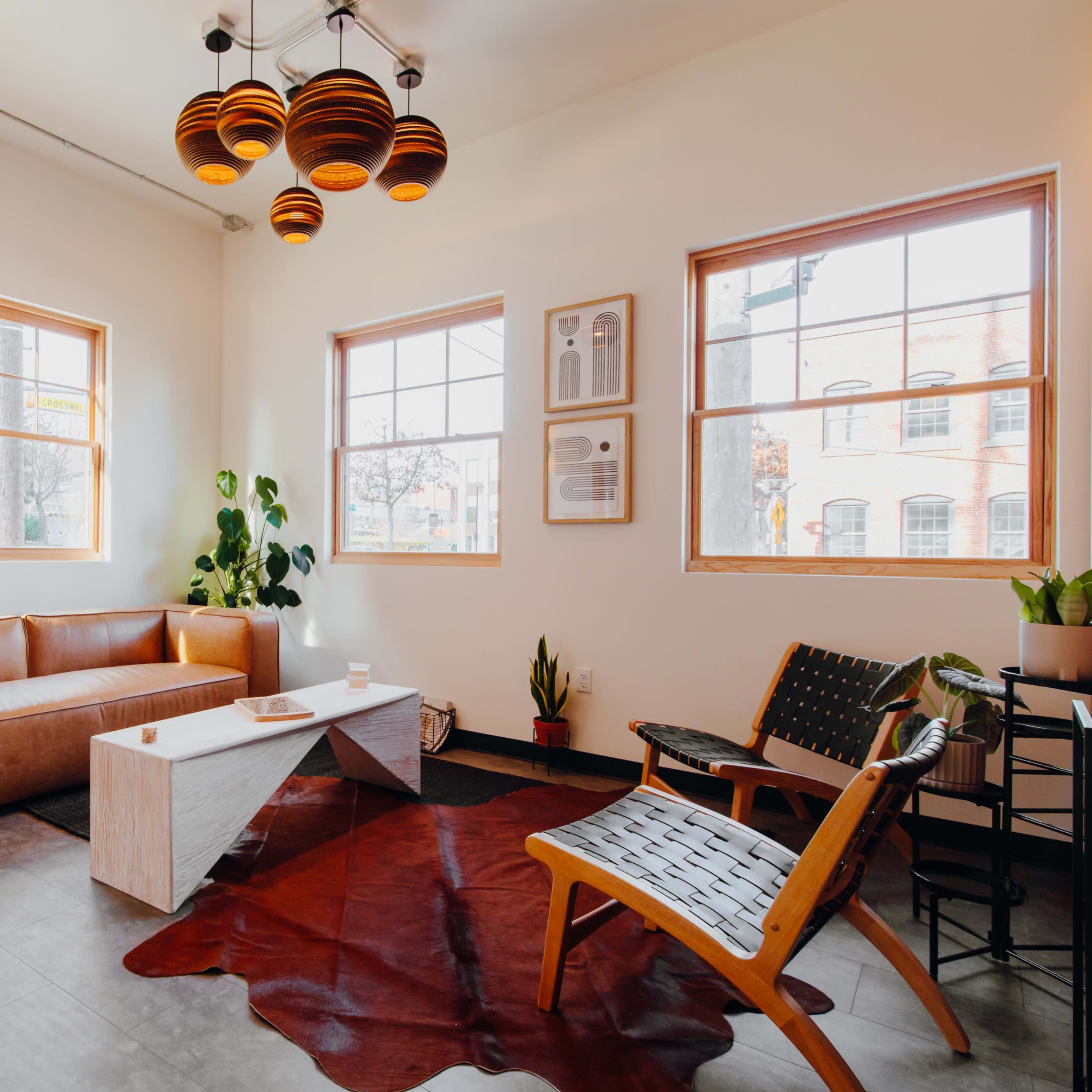 A brightly lit interior featuring a brown leather sofa, a wooden coffee table, and a unique light fixture, complemented by potted plants and large windows.