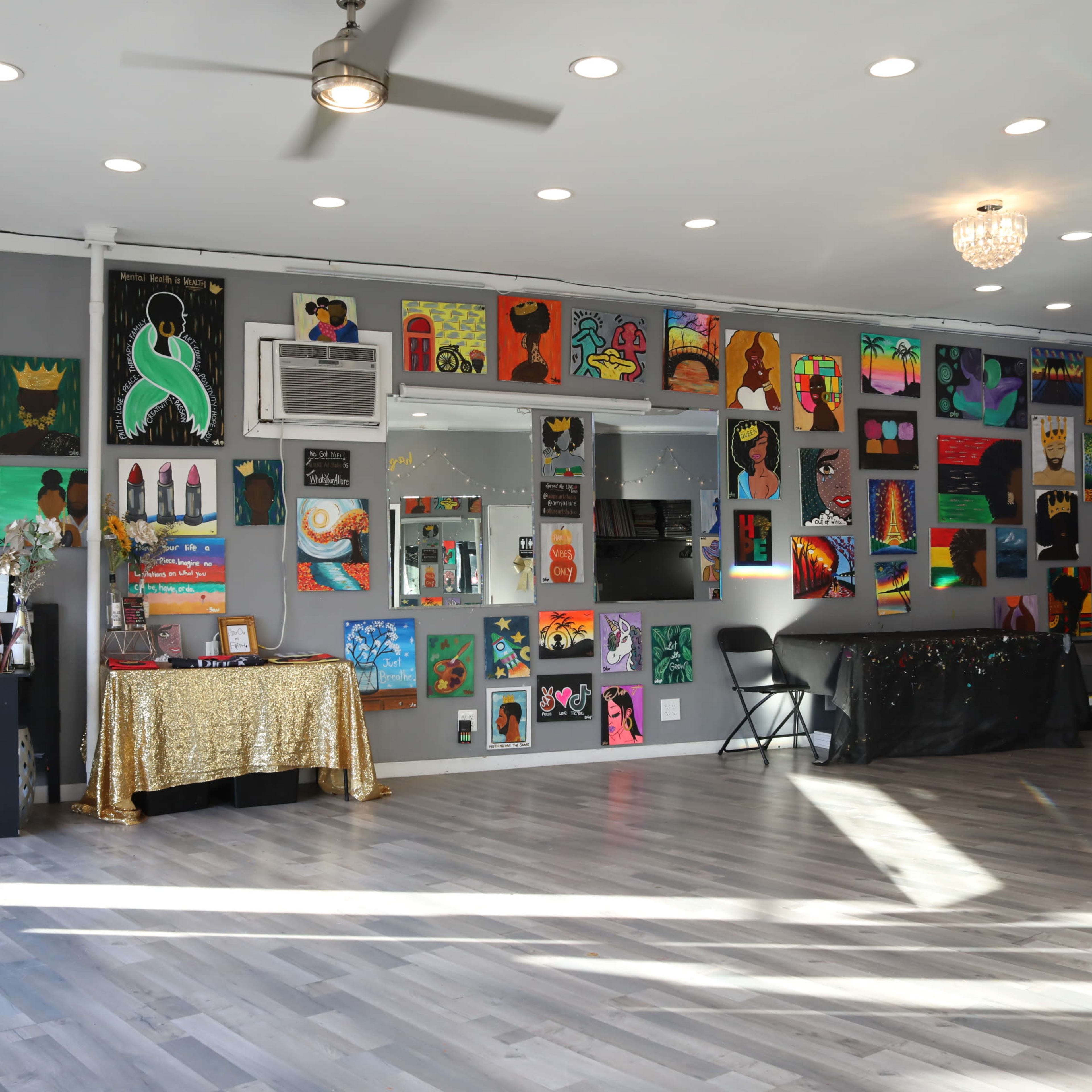 A brightly lit room with a variety of colorful paintings displayed on the walls, a small stage area, and a gathering space with chairs and a decorated table.