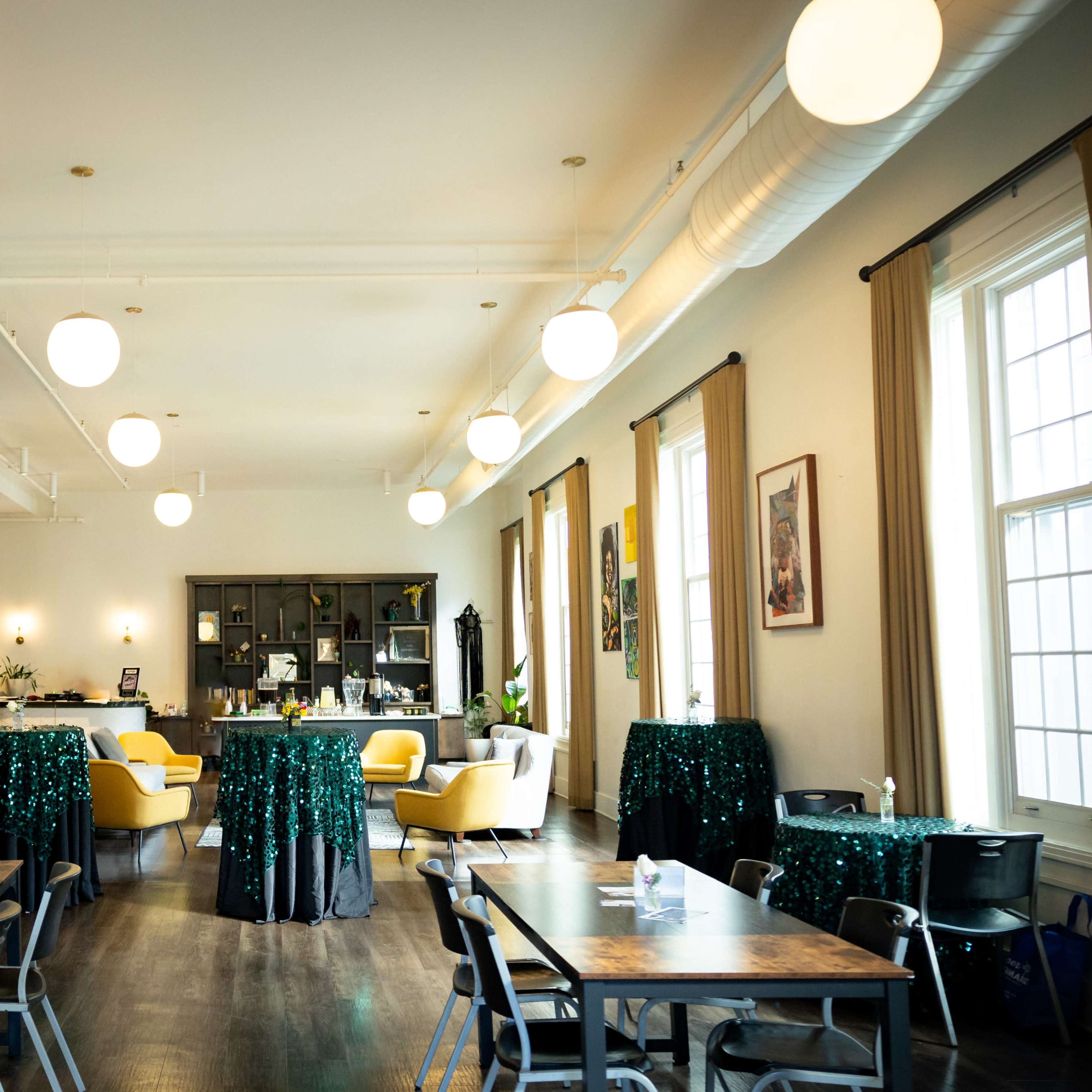 A spacious event space features tables with green sequin tablecloths and a bar area, illuminated by round pendant lights.