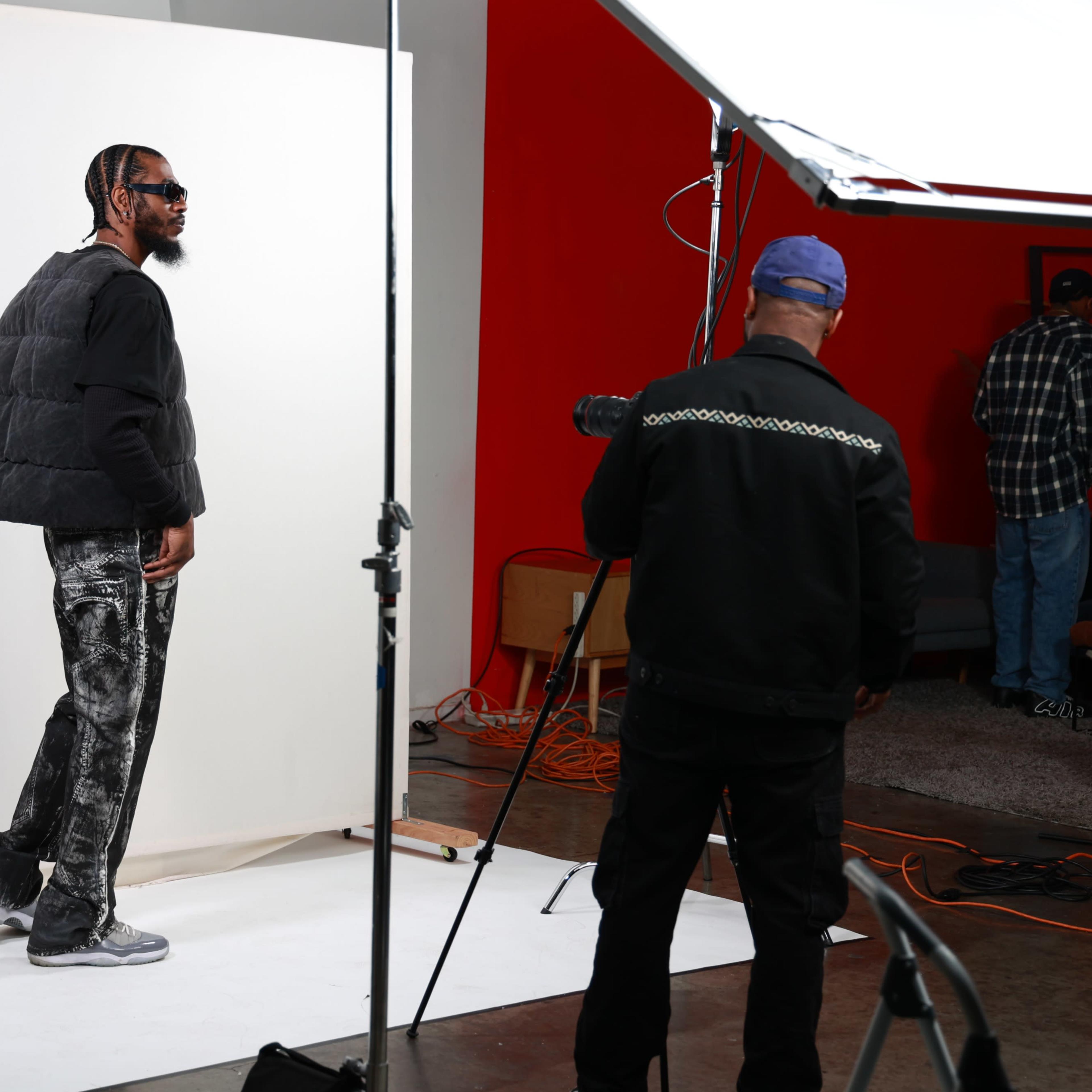 A man in a black vest poses for a photo against a backdrop while another man adjusts a camera setup in a studio.
