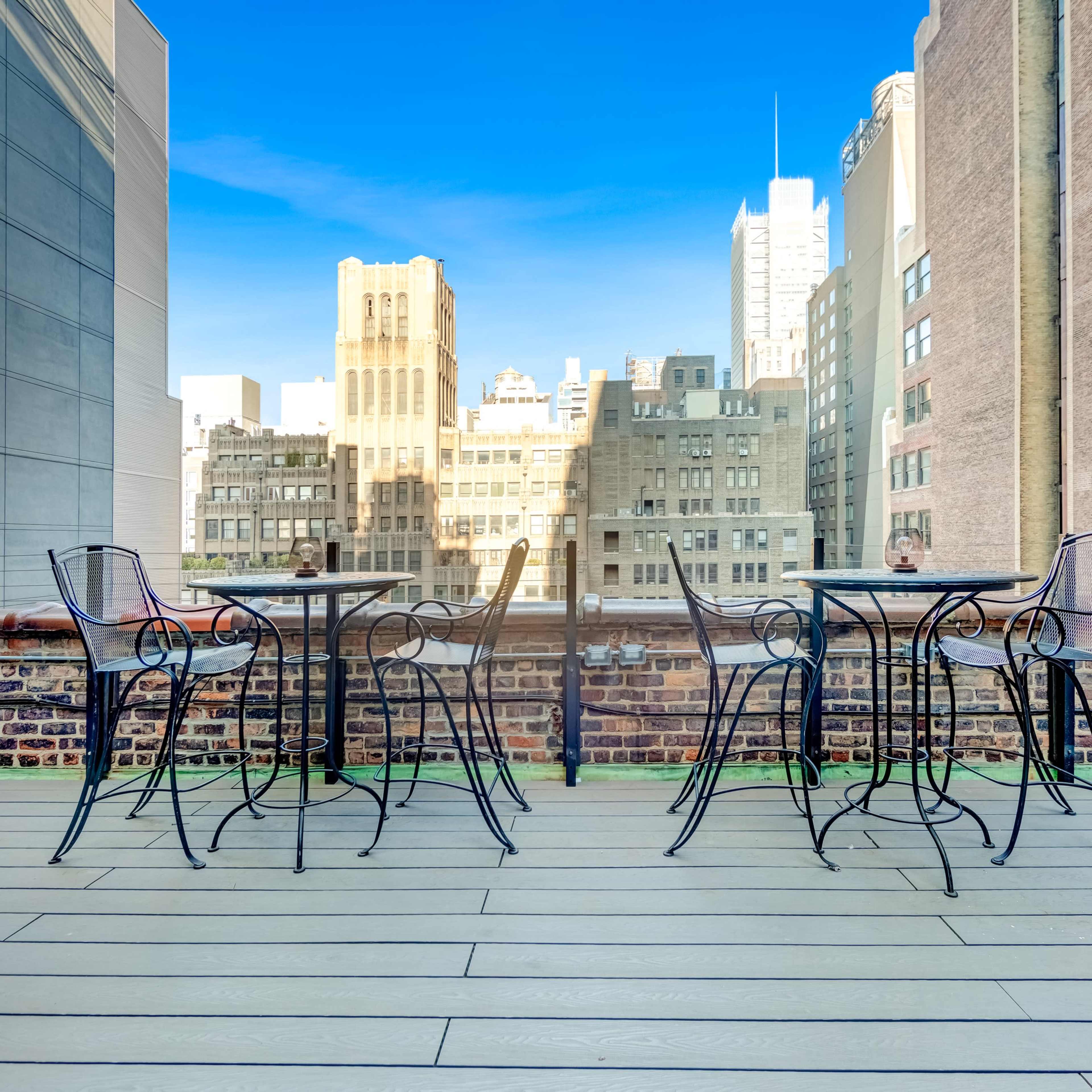 A rooftop terrace with several black metal tables and chairs overlooking a cityscape of tall buildings.