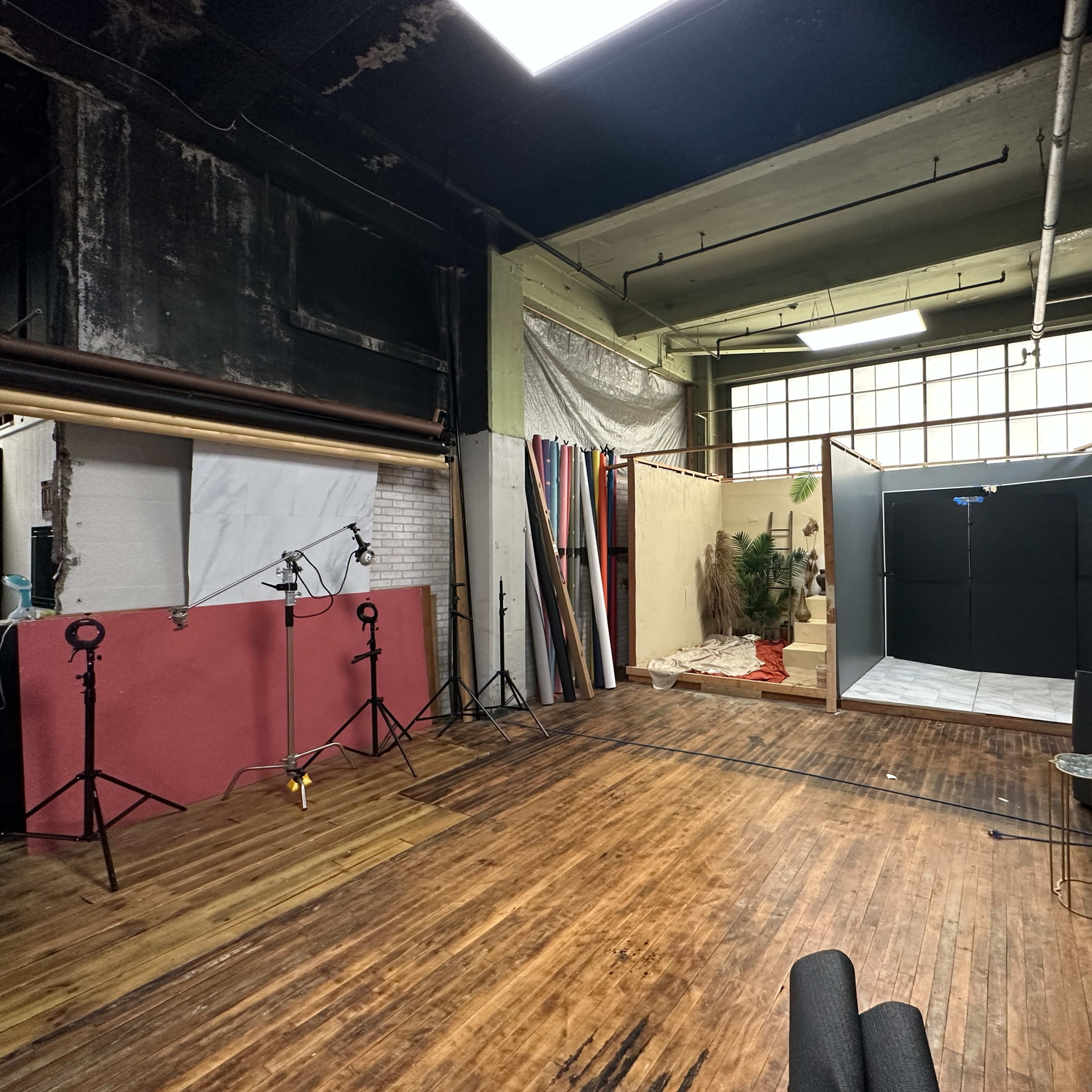 A spacious photography studio with wooden flooring, equipped with lights, a backdrop, and various props including plants and furniture.
