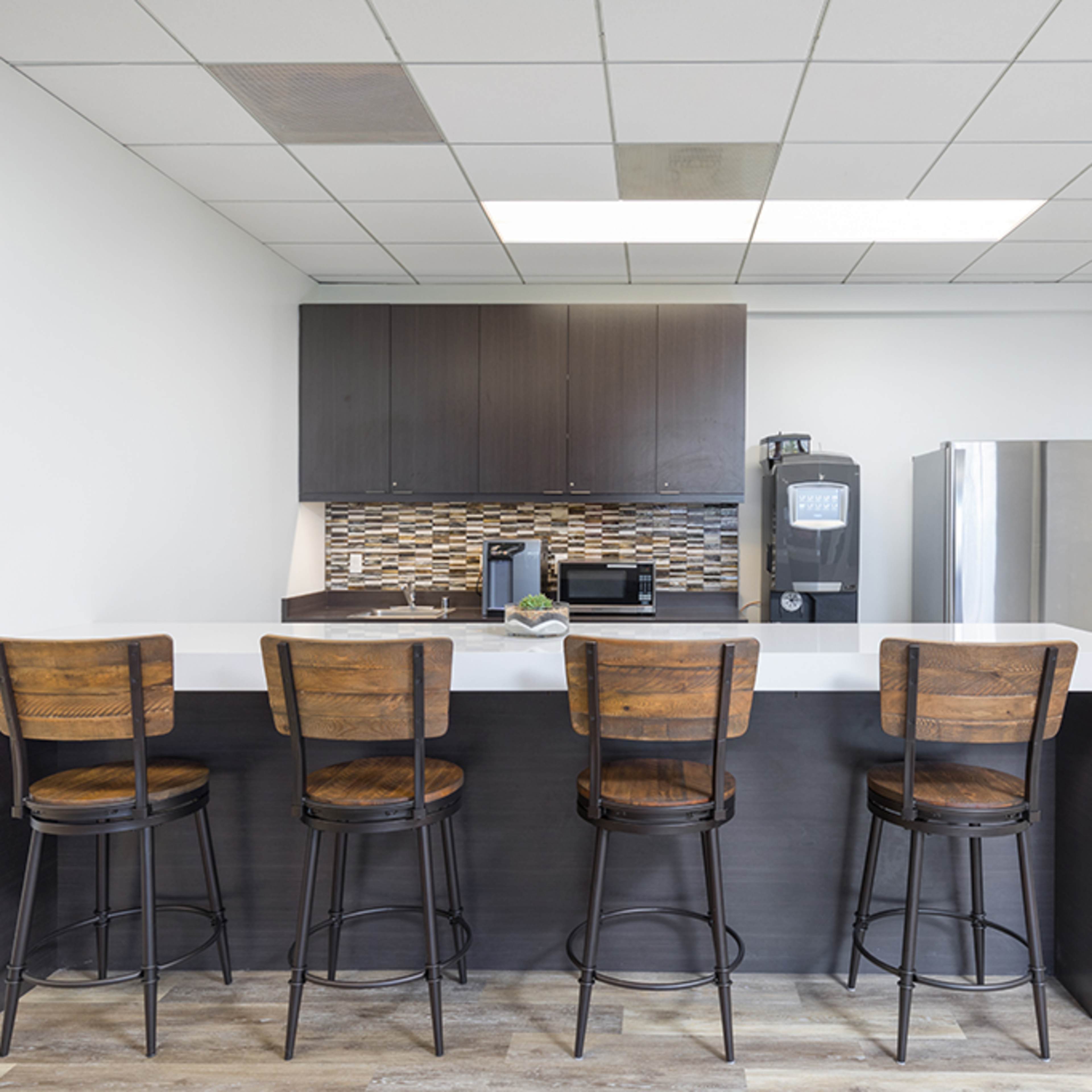 A modern kitchen features a bar with four wooden stools, a countertop, dark cabinets, a mosaic backsplash, and stainless steel appliances.