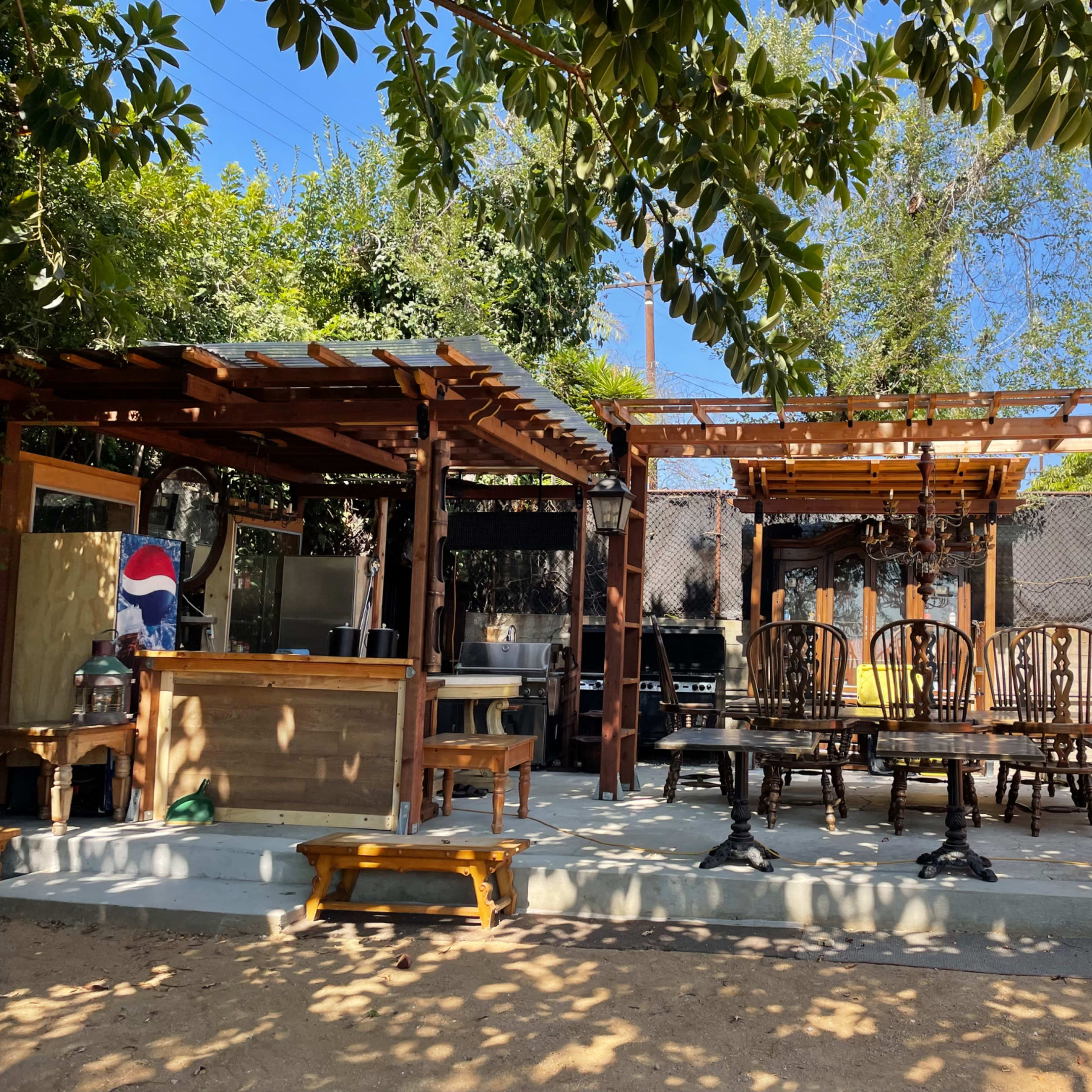 An outdoor seating area with wooden tables and chairs, a bar structure, and various trees and plants in the background.
