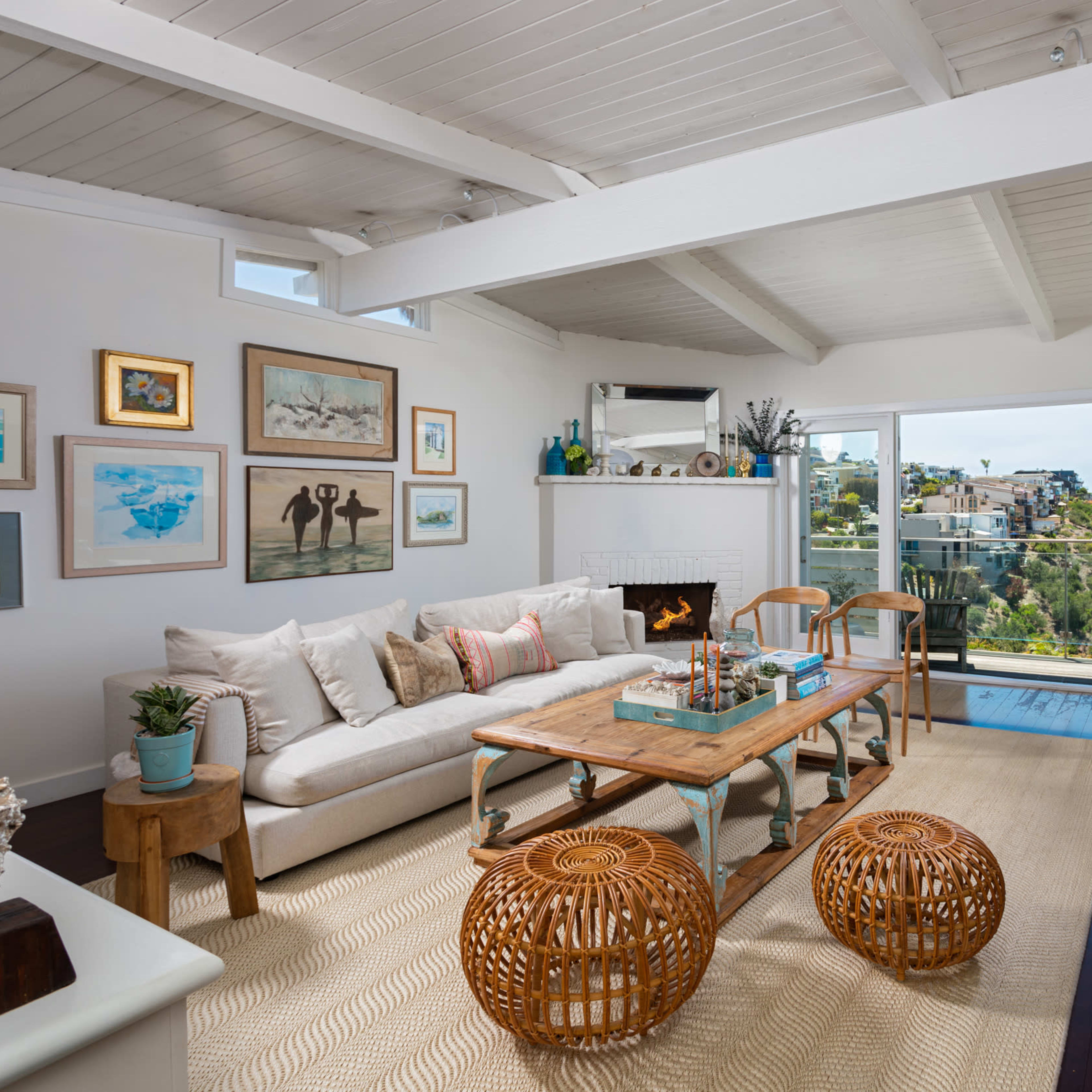 The living room features a light-filled space with a large window offering a view of the hillside, furnished with a sectional sofa, a wooden coffee table, and woven ottomans.