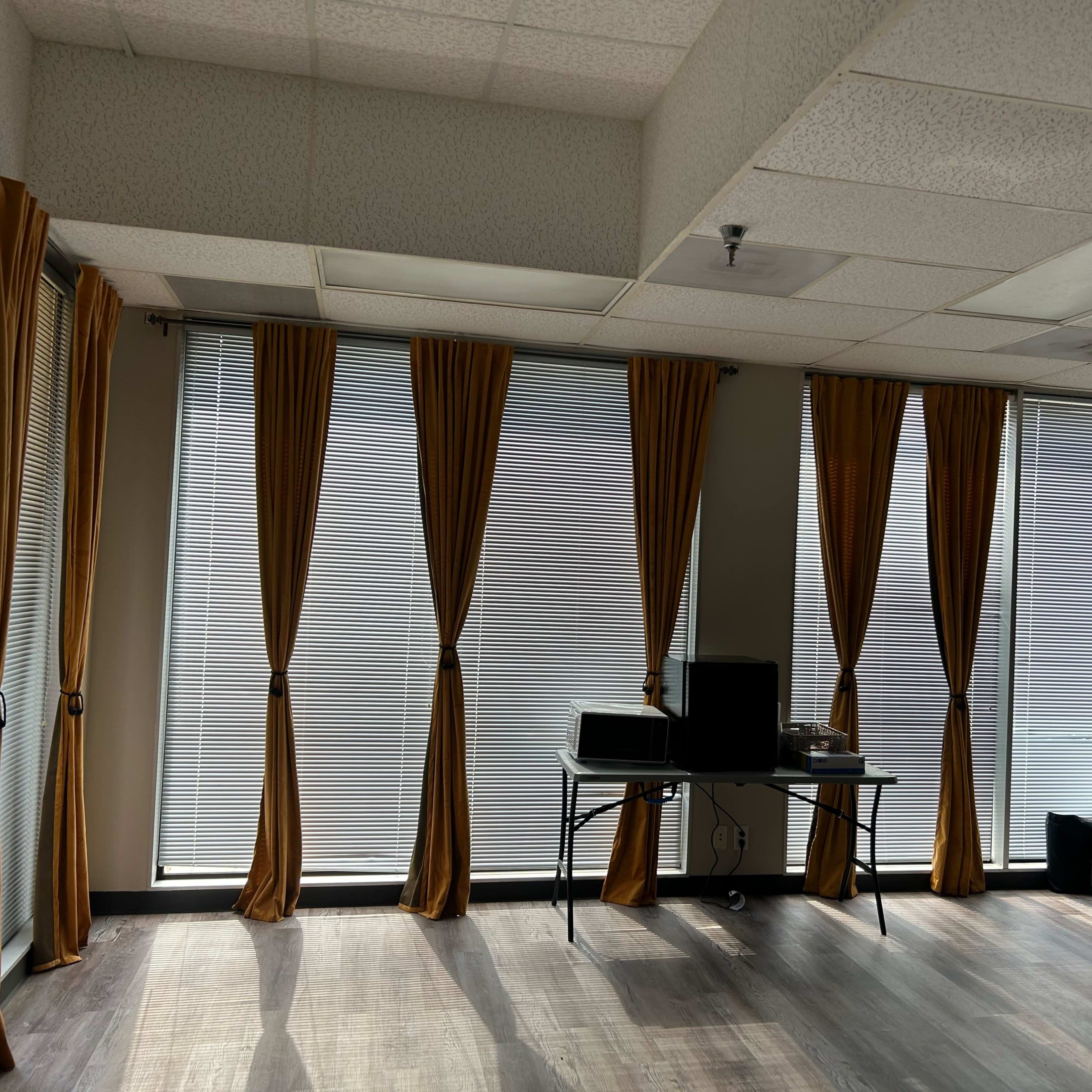 A sparsely furnished office space with large windows covered by brown curtains and a desk with a computer in front of one window.