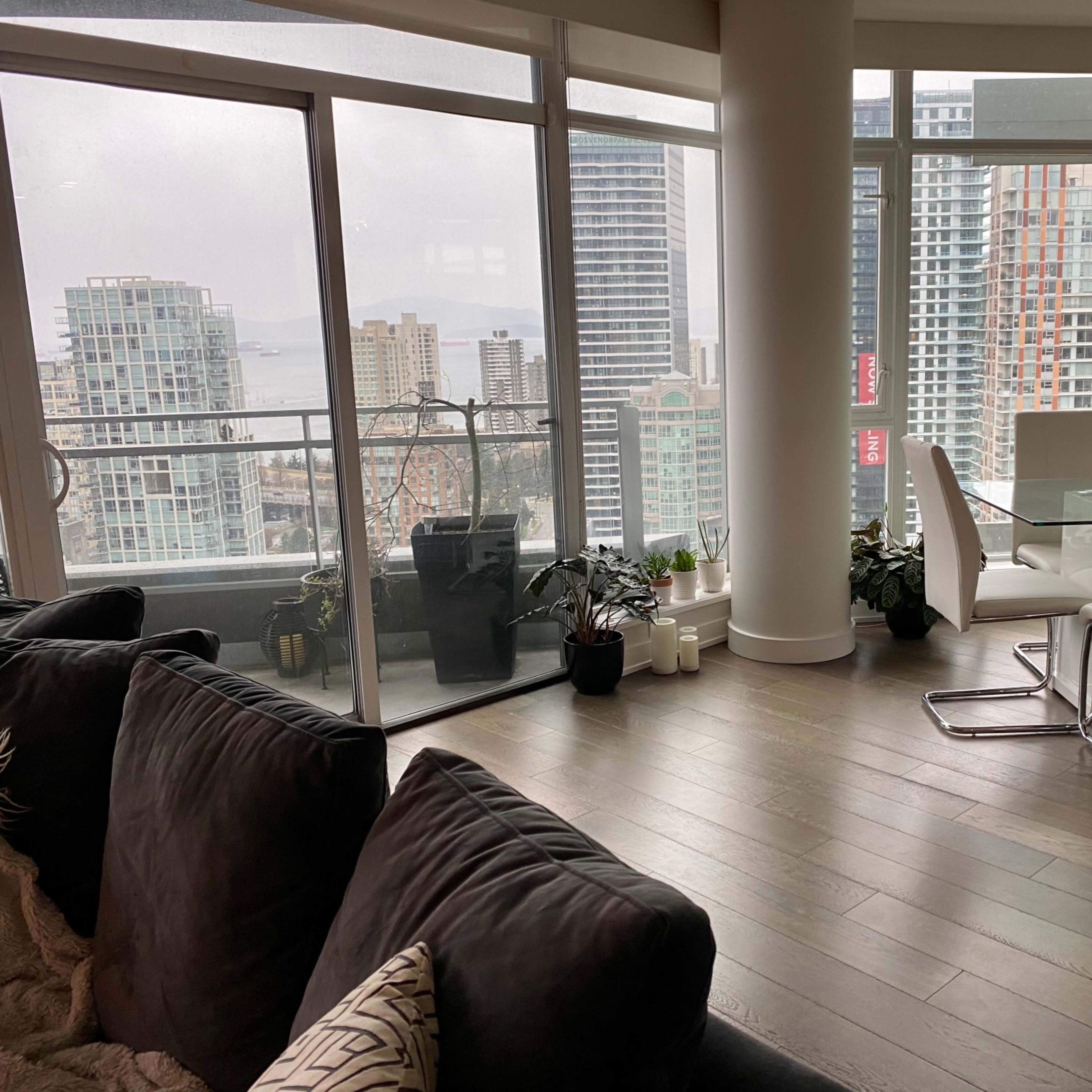 A modern apartment interior with large windows overlooking a city skyline, featuring a dining area and a cozy seating arrangement.