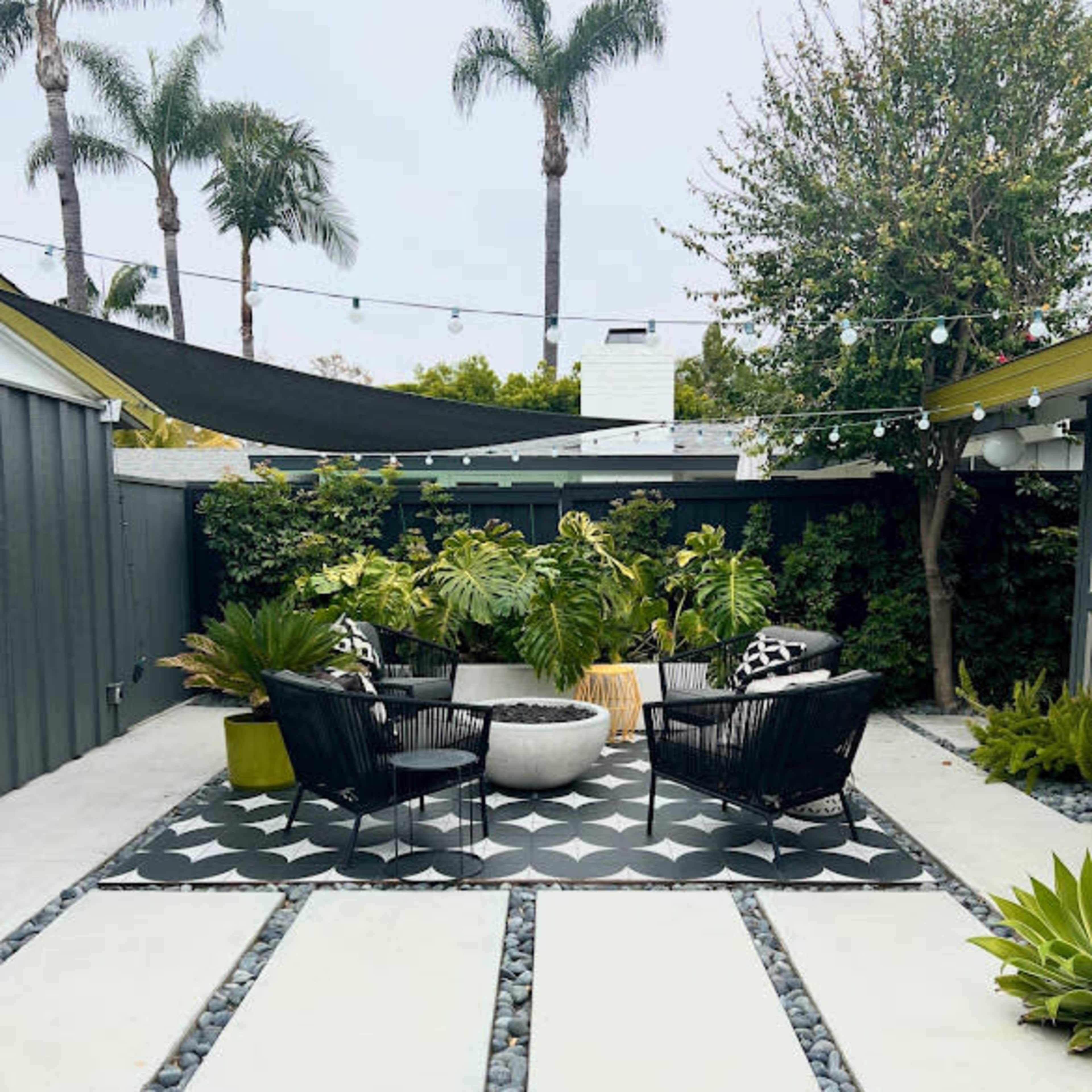 A modern outdoor patio features two black wicker chairs facing a round concrete table, surrounded by lush greenery and decorative stones.