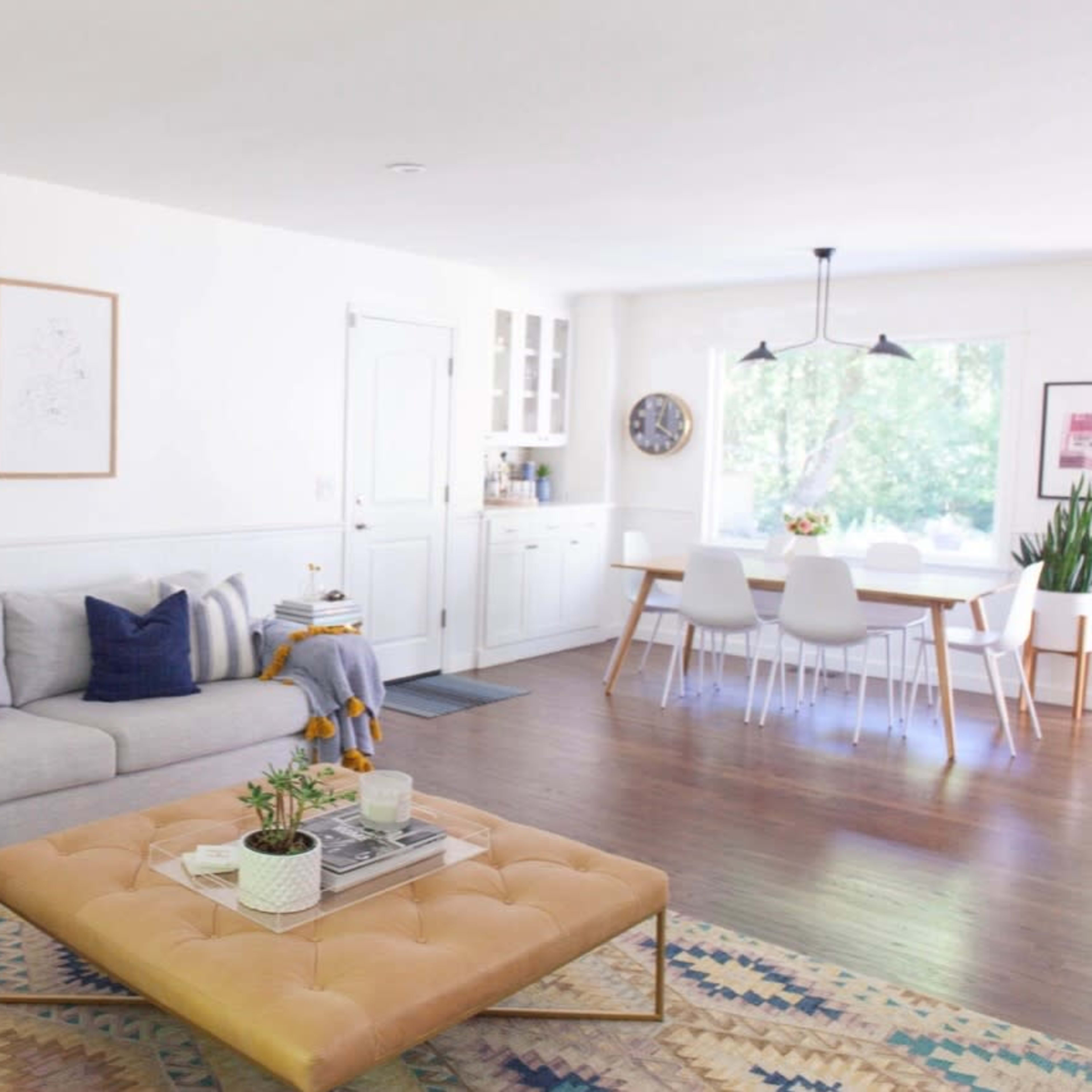 A bright living room with a gray sofa, a large tan ottoman, a patterned rug, and a dining area with white chairs and a large window.