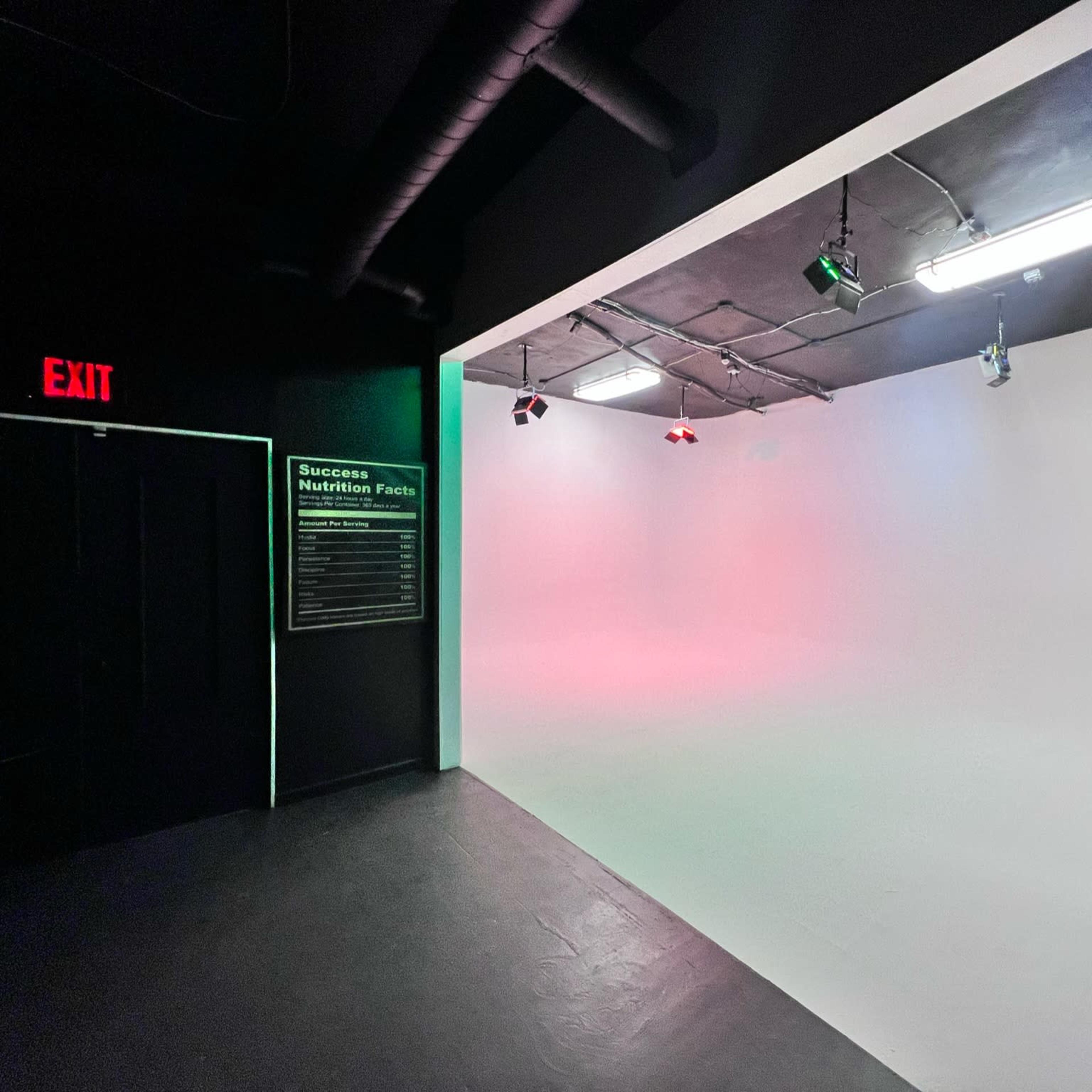 A studio space with a plain white backdrop, lighting fixtures on the ceiling, and an exit sign on the left wall.