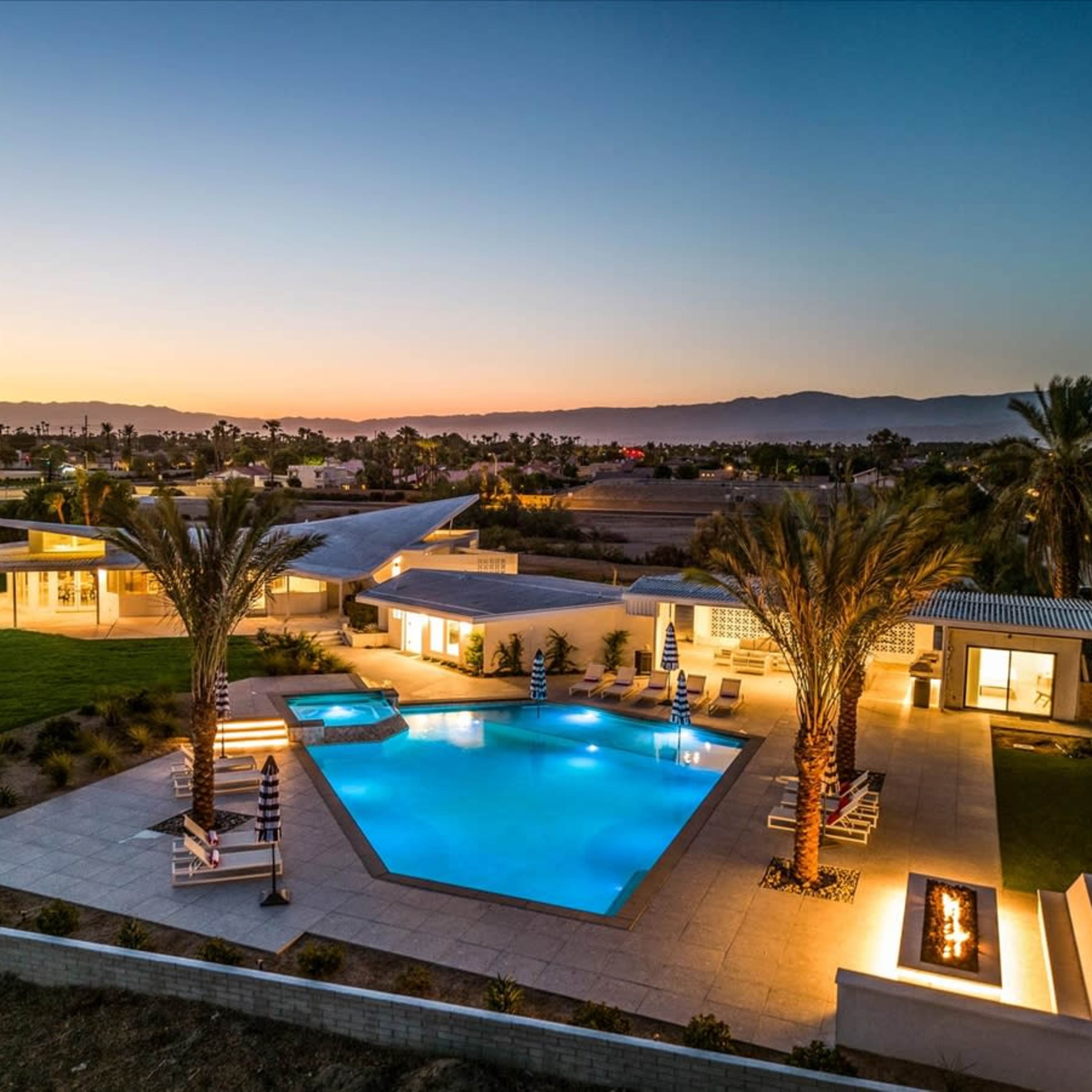 A modern house with a pool and palm trees, illuminated by soft lights against a sunset backdrop.