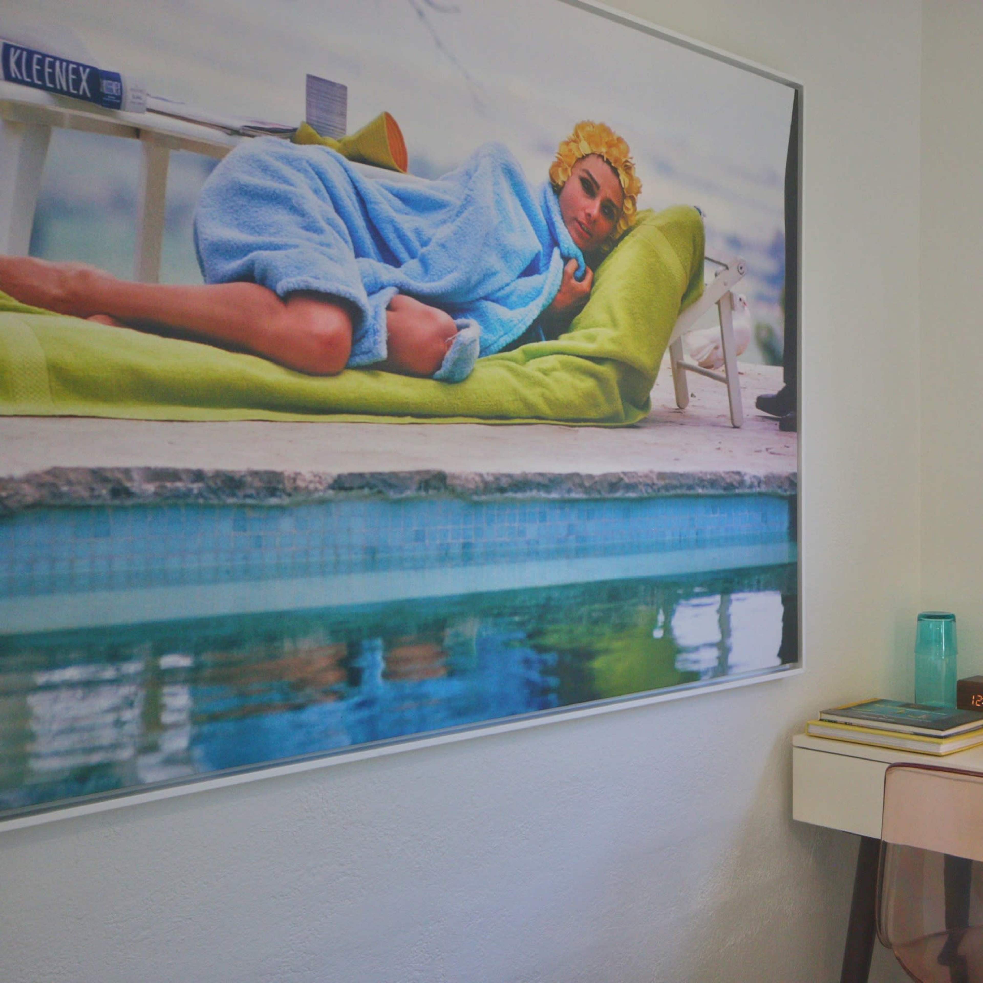 A modern room featuring a large framed artwork of a person lounging on a green sofa next to a pool, with a desk, laptop, and bedside lamp nearby.