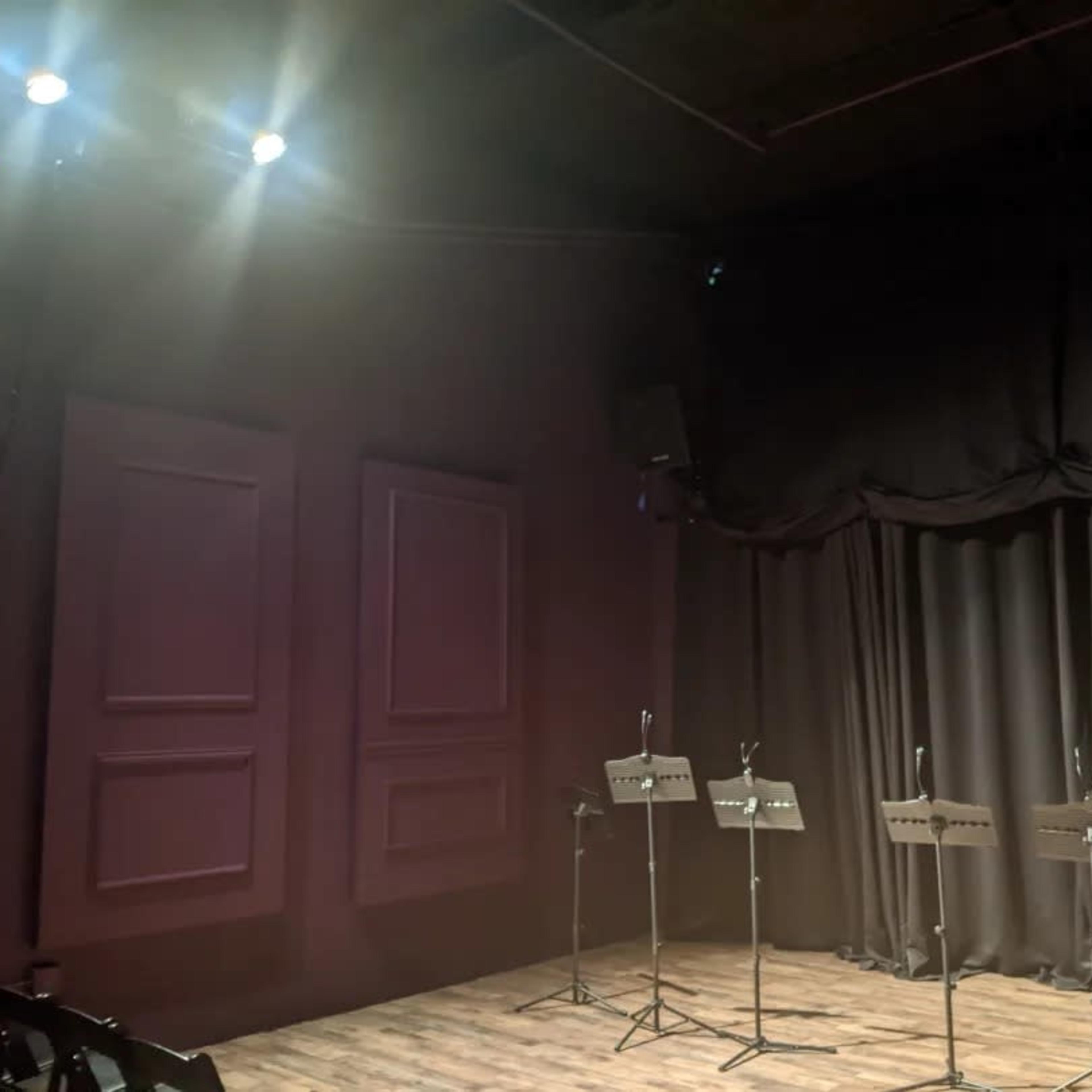 A dimly lit performance space with black curtains and several music stands arranged in front of them.