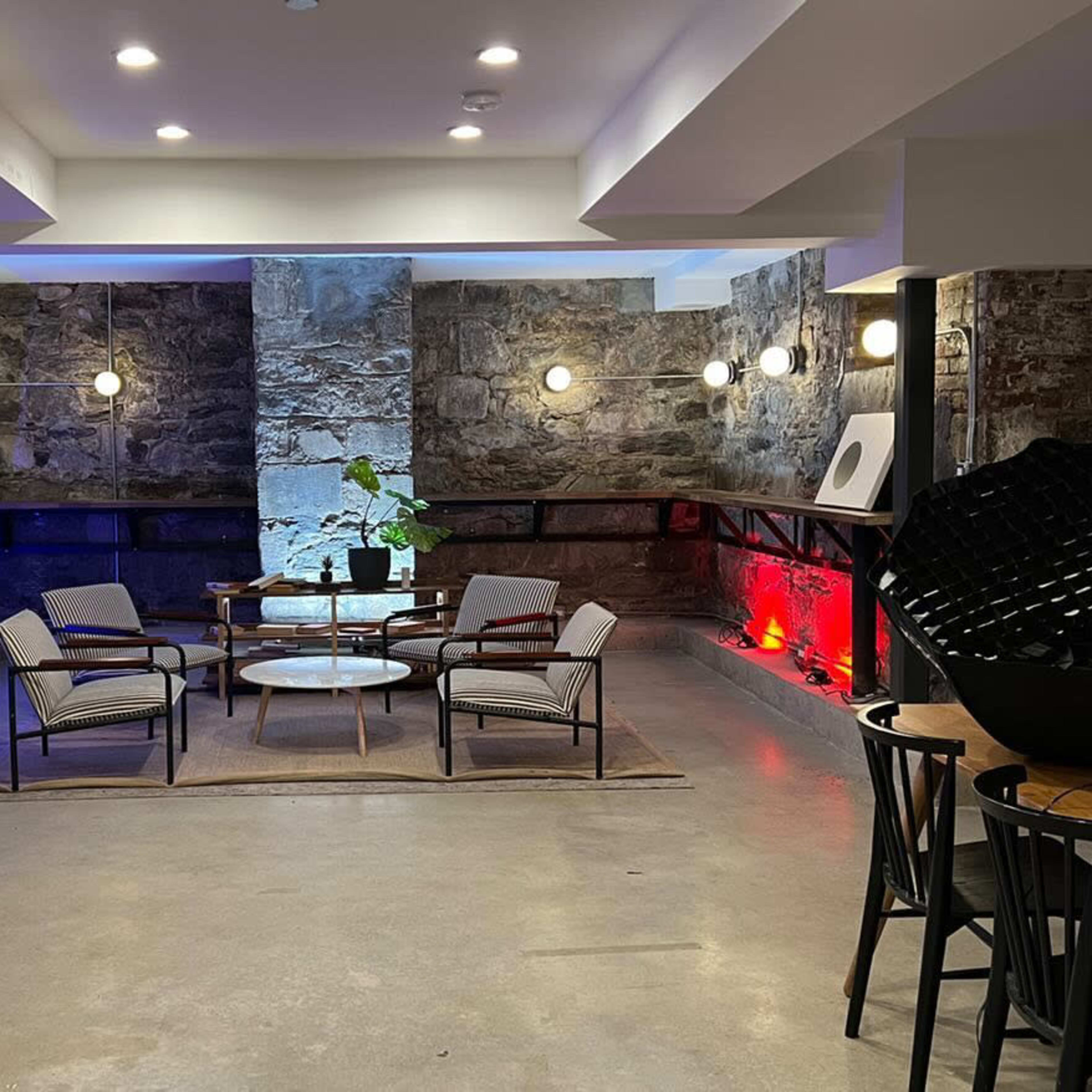 A modern seating area with four chairs and a glass table is illuminated by wall-mounted lights and colorful accents against a stone wall backdrop.