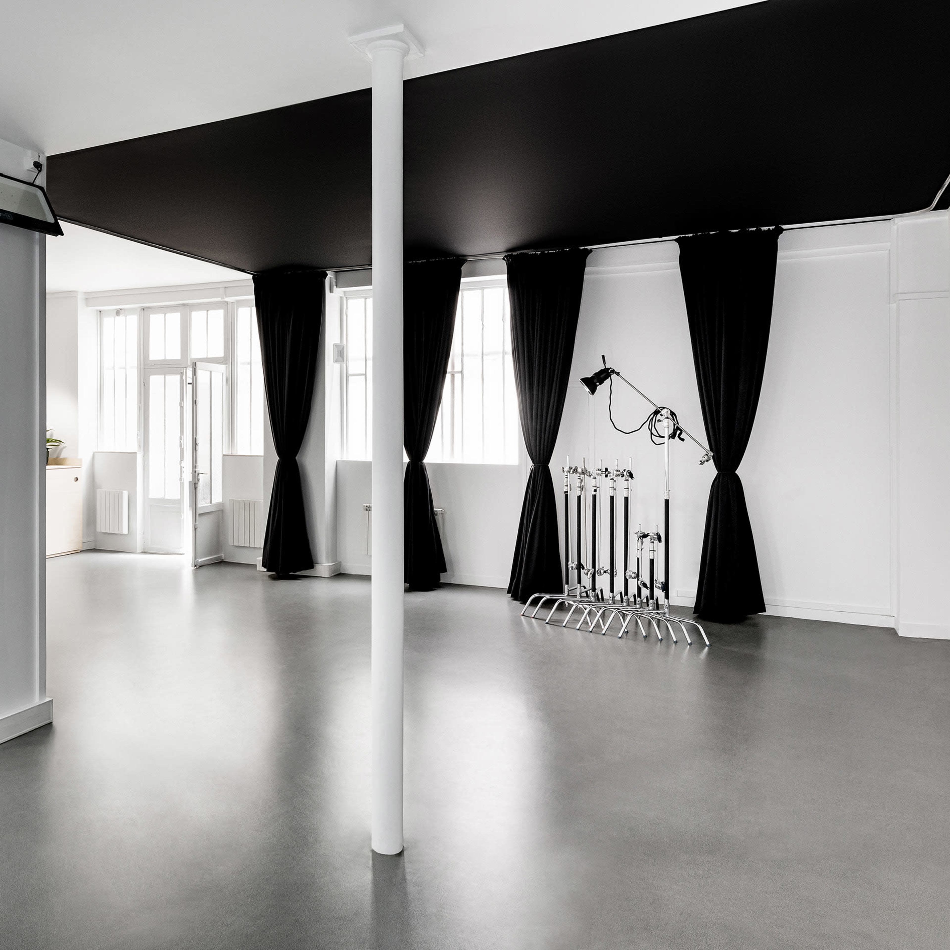 A spacious, minimalistic studio features white walls, a black ceiling, large windows, and a curtained area with equipment stands.