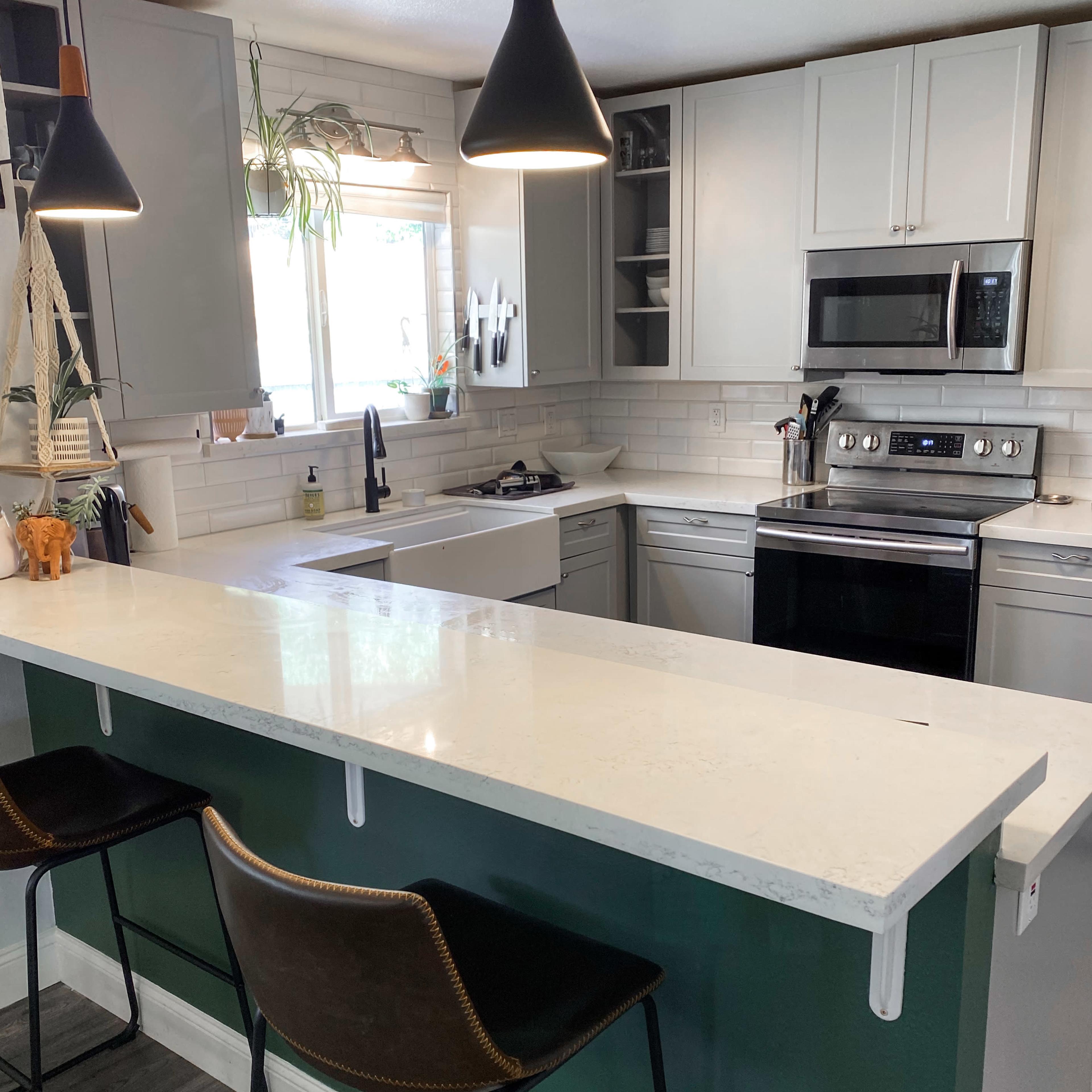 A modern kitchen features a white countertop, stainless steel appliances, and dark pendant lights above a bar with two metal stools.