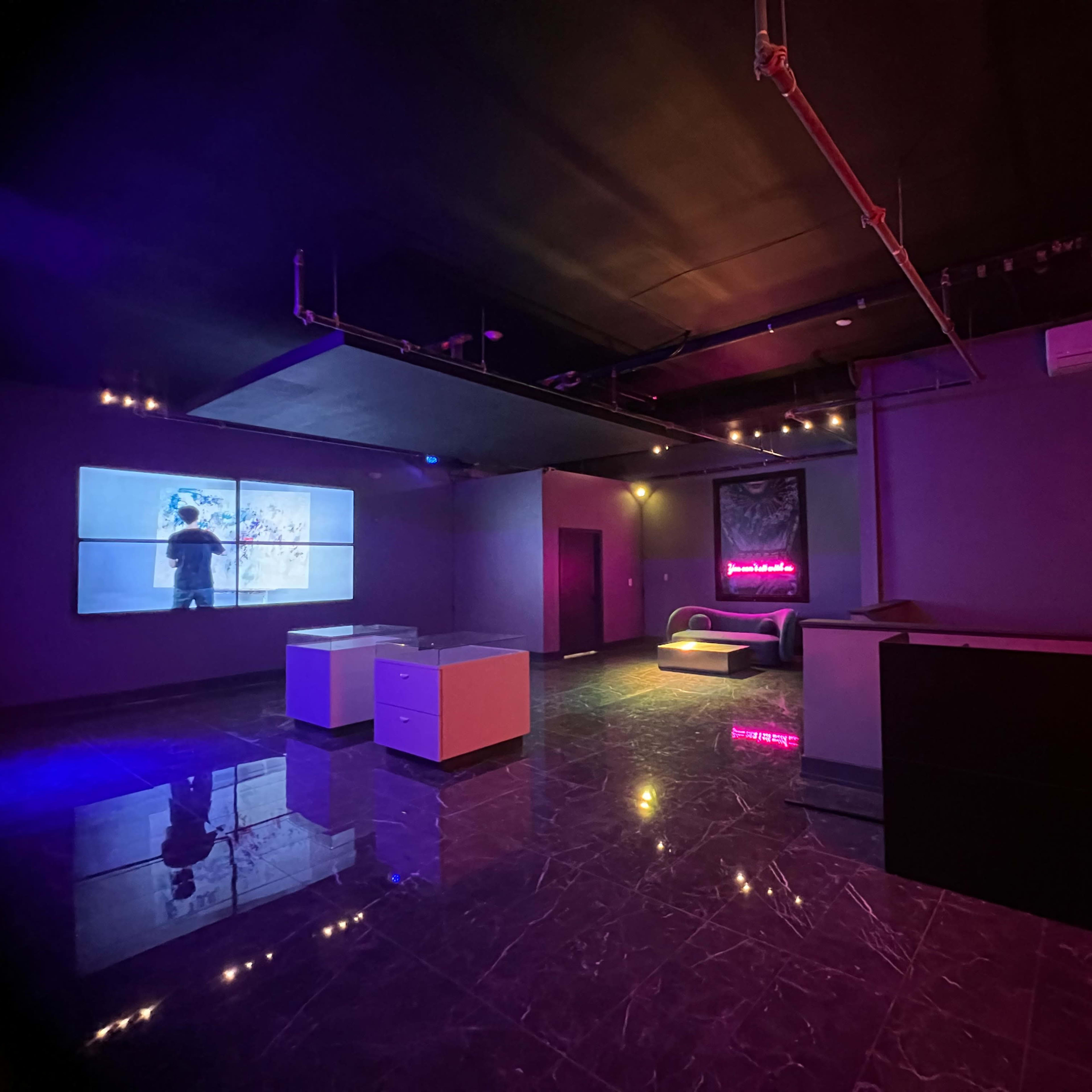 The interior of a dimly lit room features a large screen displaying a figure, modern furniture, and colorful lighting effects.