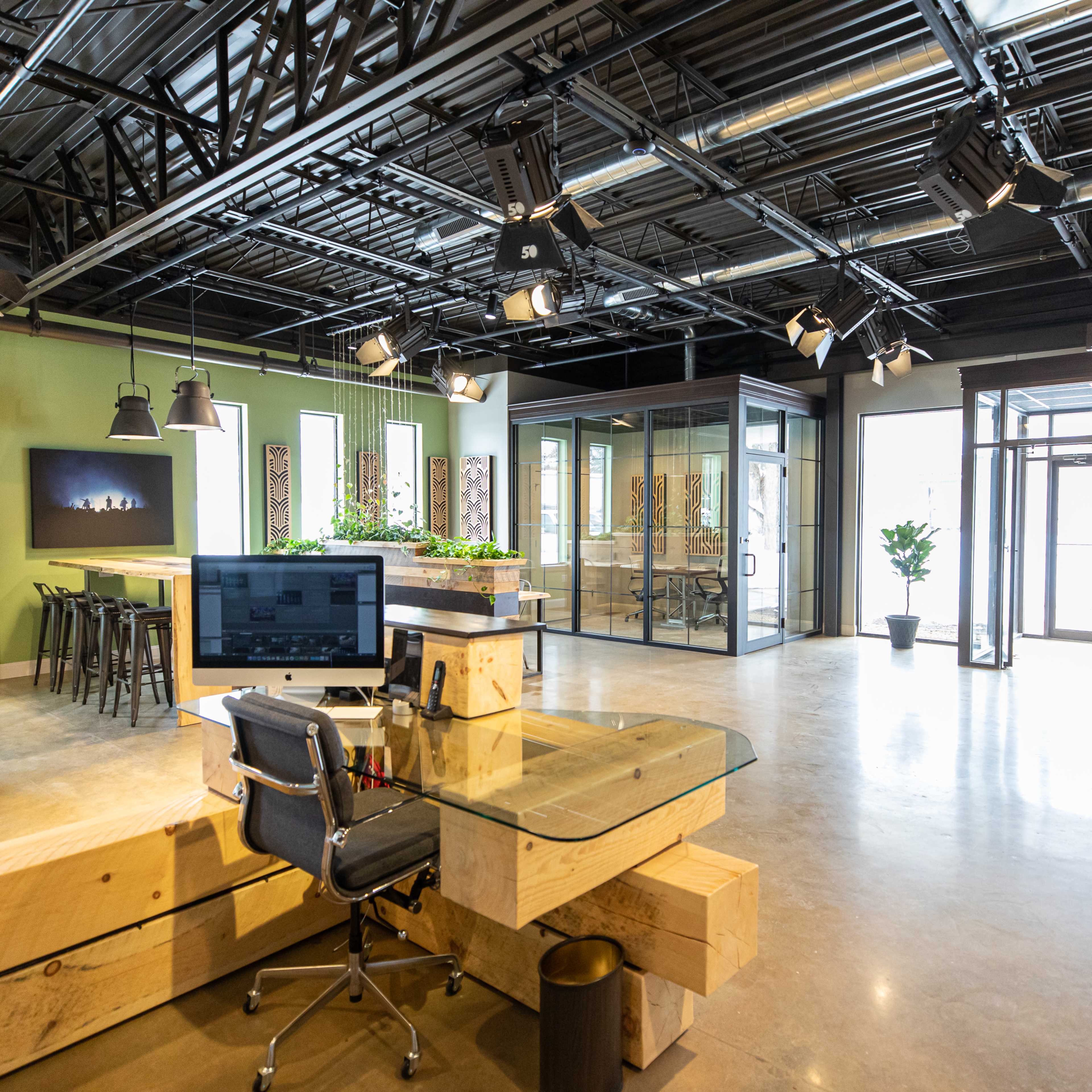 A modern office space featuring a desk with a computer, a seating area with bar stools, and glass-walled meeting rooms.