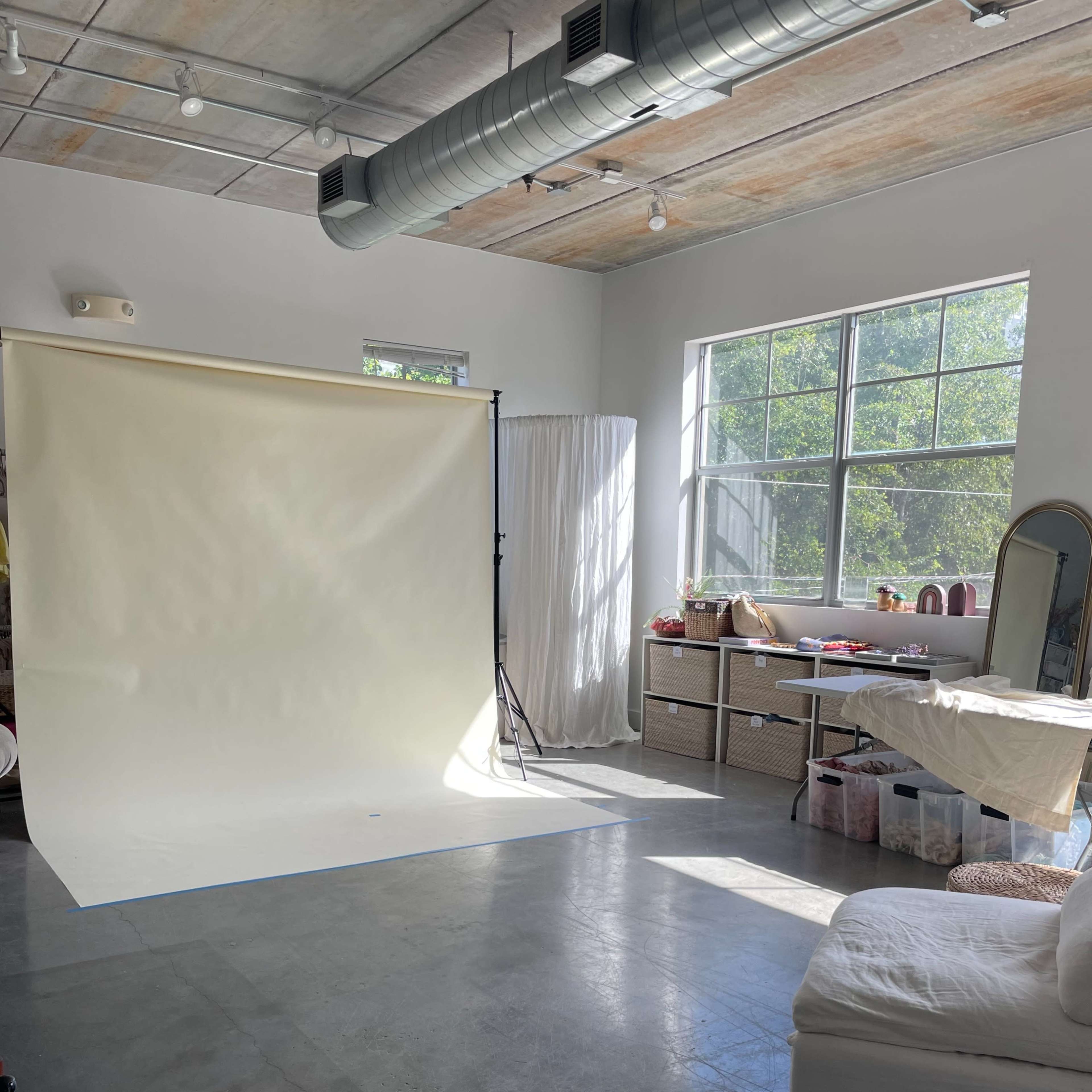 A well-lit photography studio with a neutral backdrop, large windows, and various furniture pieces and props arranged around the space.