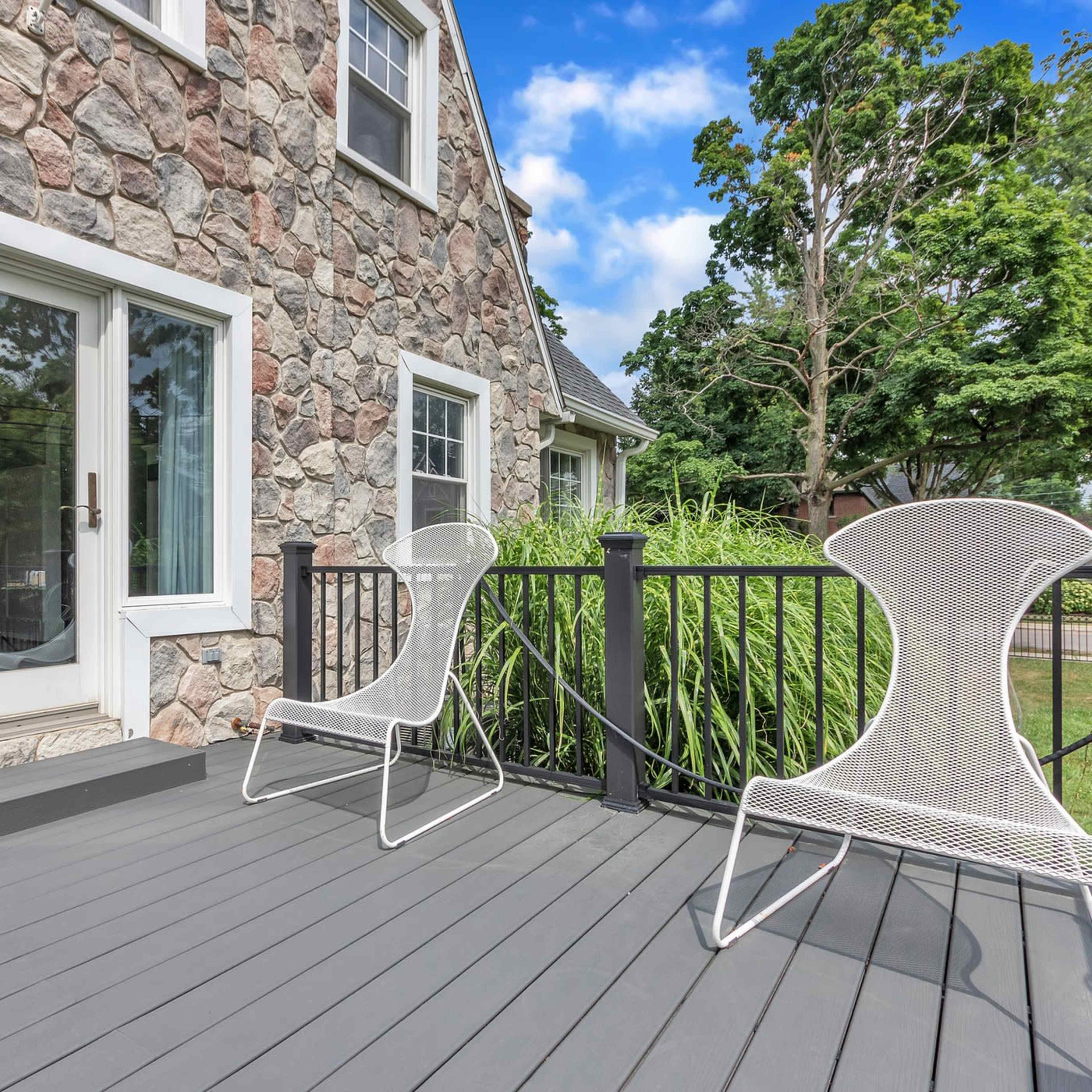 A stone house features a wooden deck with two white wire chairs and greenery in the background.