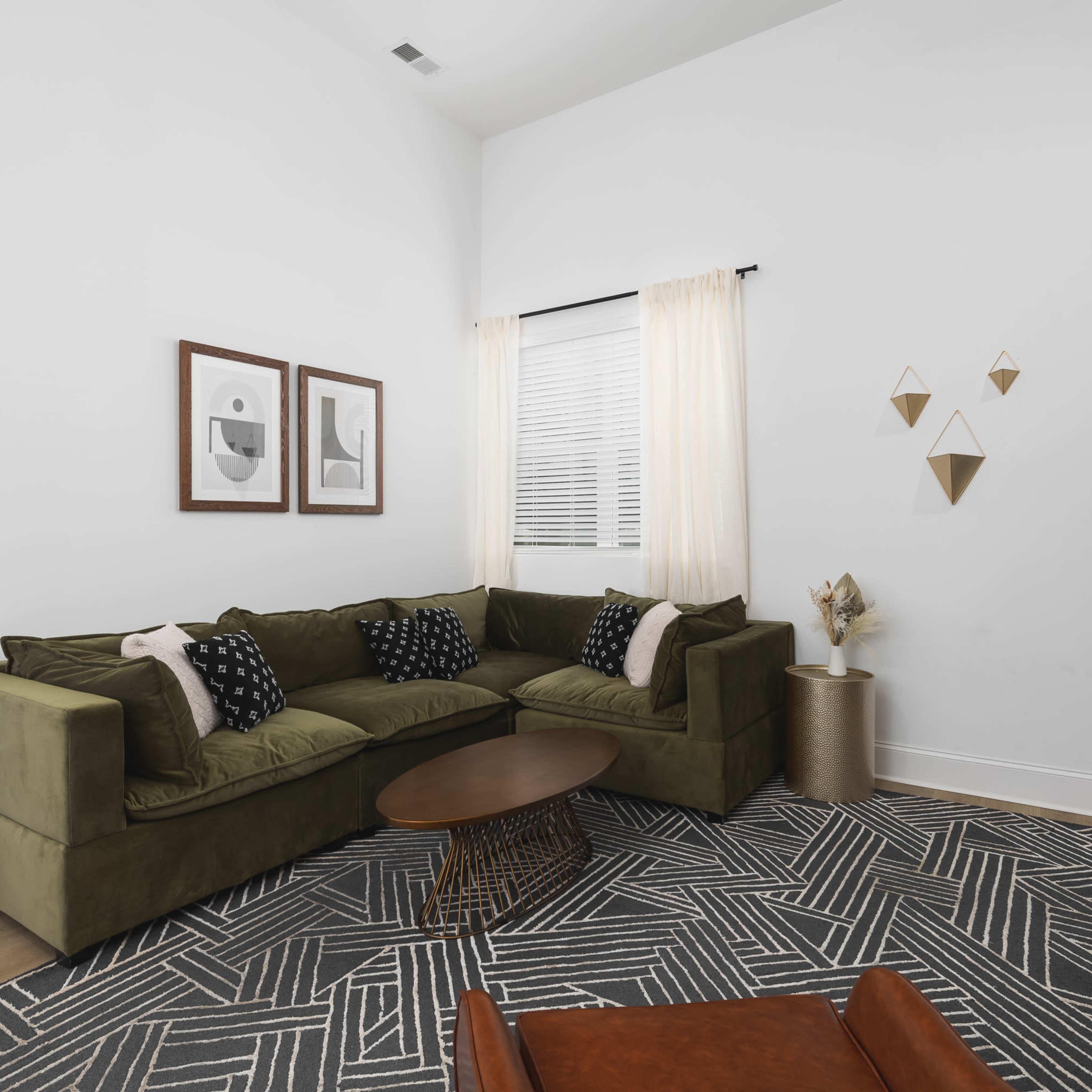 A green sectional sofa is arranged in a bright living room featuring a round coffee table, two framed artworks on the wall, and decorative items on a shelf.